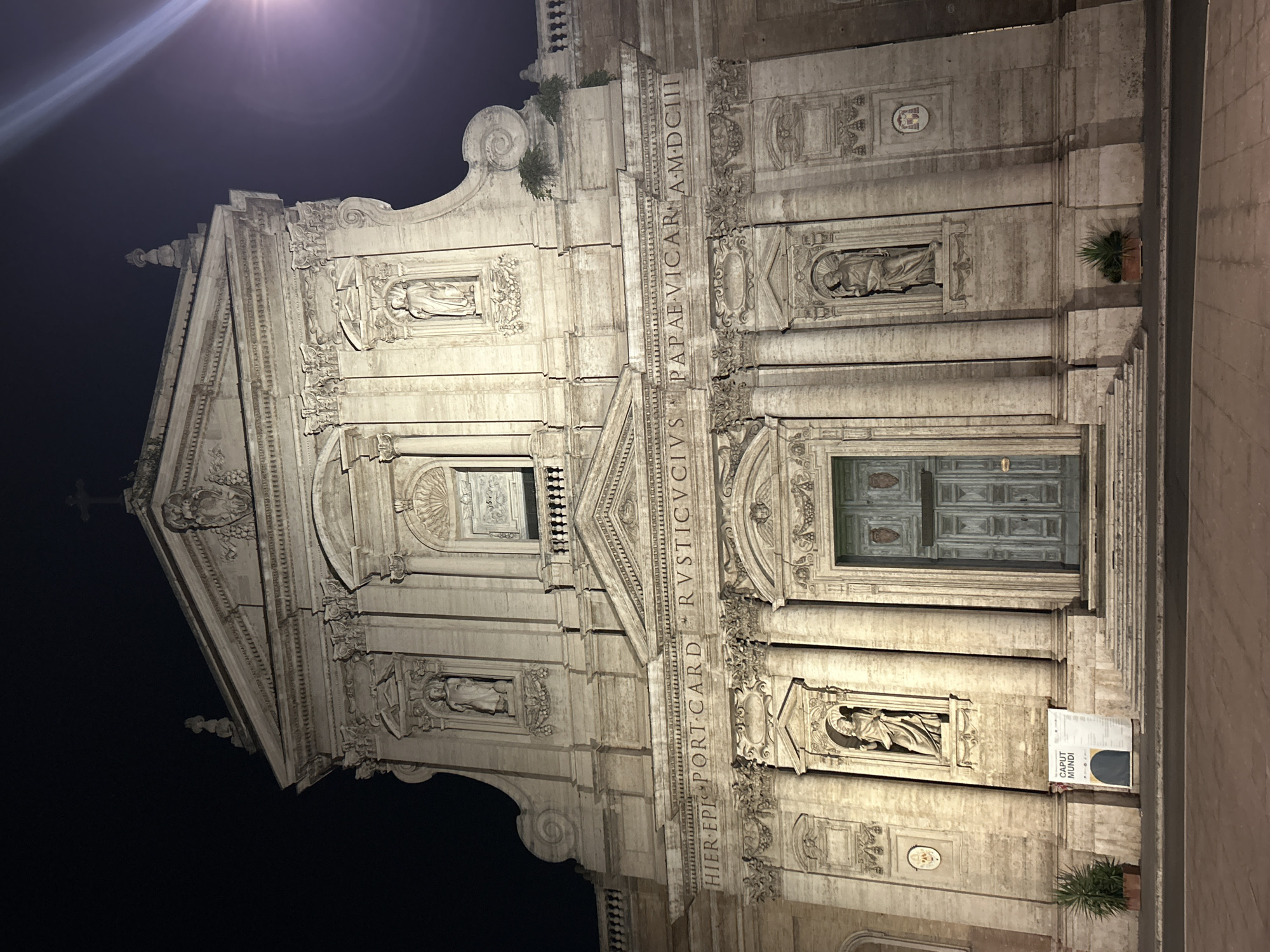 A night-time, eye-level, front-facing exterior shot shows the facade of the Santa Maria Maggiore Basilica in Rome, Italy. The basilica is illuminated by artificial lights, casting a warm glow on its travertine stone structure. The facade features a large, arched wooden double door in the center, flanked by two statues in niches. Above the door, an inscription reads "HIER EPIS PORT CARD RVSTICVCIVS PAPAE VICAR A MDCIII". Further up, the facade is adorned with more statues in niches, intricate carvings, and architectural elements characteristic of Baroque style. The sky is dark, suggesting it is late at night. Two potted plants are visible in front of the basilica, one on the left and one on the right. A large banner with the text "CAPUT MUNDI" is displayed to the left of the entrance. The ground in front of the basilica is paved with stone. The overall scene is grand and imposing, conveying a sense of historical and religious significance.