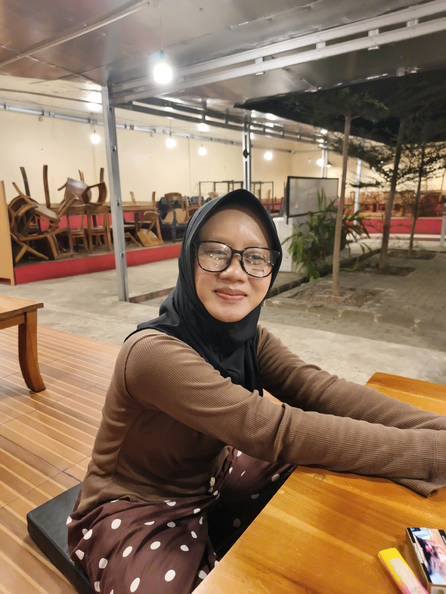A medium shot shows a young woman wearing a black hijab and a brown long-sleeved shirt with white polka dot pants. She is sitting at a wooden table, looking at the camera and smiling. She wears black-rimmed glasses that say "MOLSION" on the side. In the background, chairs are stacked on tables in what appears to be an outdoor dining area. The scene is lit by overhead lights and appears to be located in Ketapang, Indonesia.
