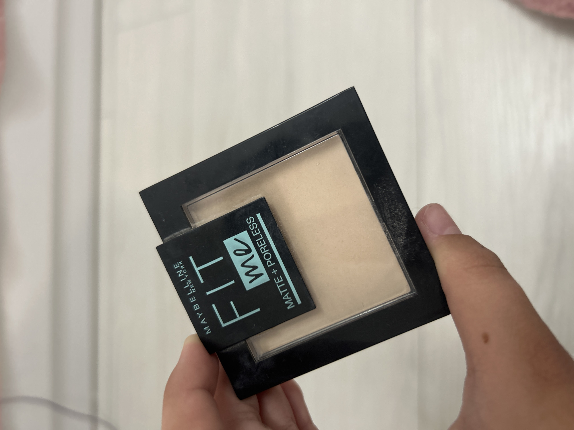 A hand is holding a black compact of Maybelline Fit Me Matte + Poreless powder. The powder inside is a light beige shade. The compact is sitting on a light-colored, textured surface, possibly a table or floor, with faint horizontal lines visible. The background is out of focus and appears to be a wall or similar flat surface. There are no other people or objects clearly visible in the image. The lighting is neutral, with no harsh shadows.