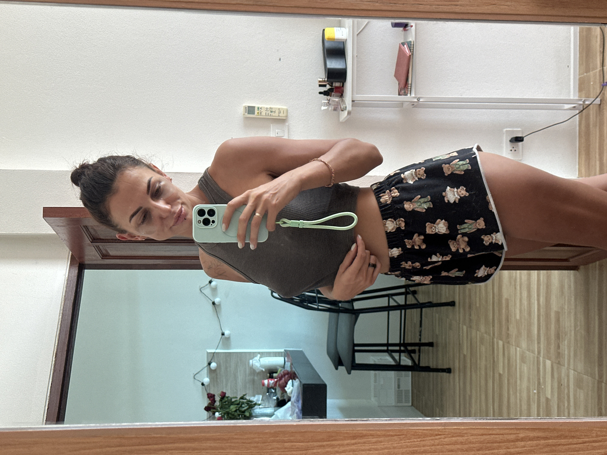 A woman is standing in what appears to be a hotel room or apartment in Ko Samui District, Thailand, taking a mirror selfie. She is wearing a dark grey crop top and black shorts with a teddy bear pattern. Her hair is pulled up in a bun. The room has a wooden door frame, a mirror, a counter with some items on it, and a white shelf with various objects. A string of lights is visible in the reflection. The flooring is wood-look tile.