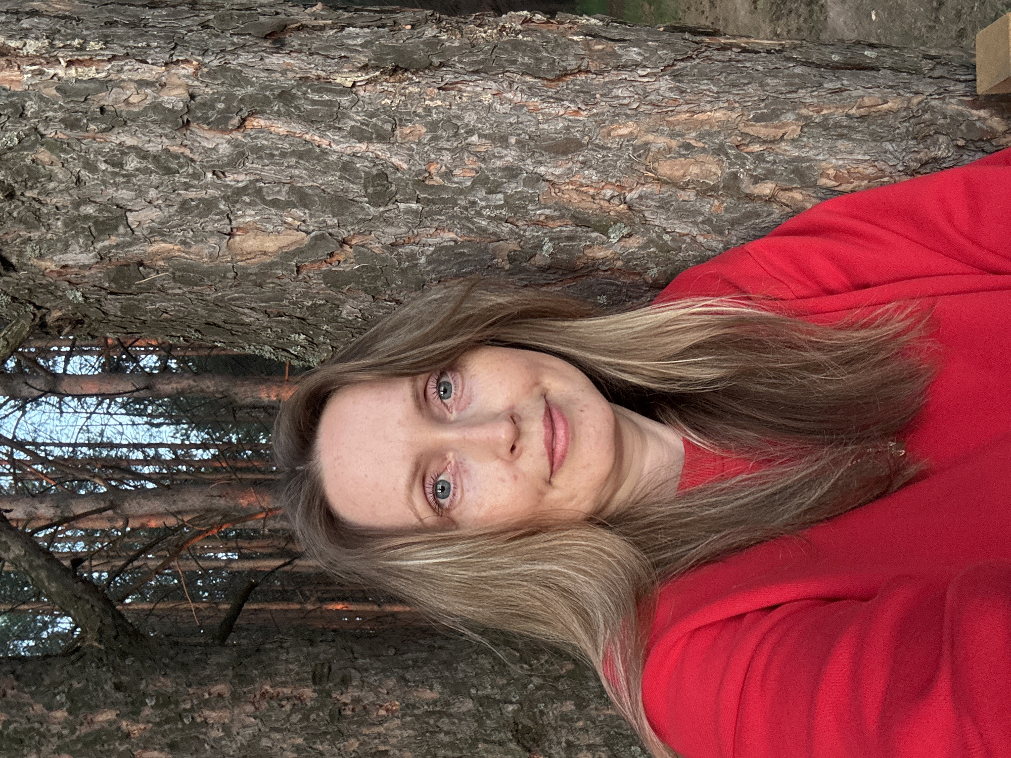 A woman with shoulder-length blonde hair and blue eyes, wearing a red sweater, is pictured outdoors, leaning against a large tree trunk. The background consists of a dense forest with tall trees. The lighting suggests it is either dawn or dusk, as there is a warm, orange glow on some of the tree trunks. The overall scene is natural and serene.
