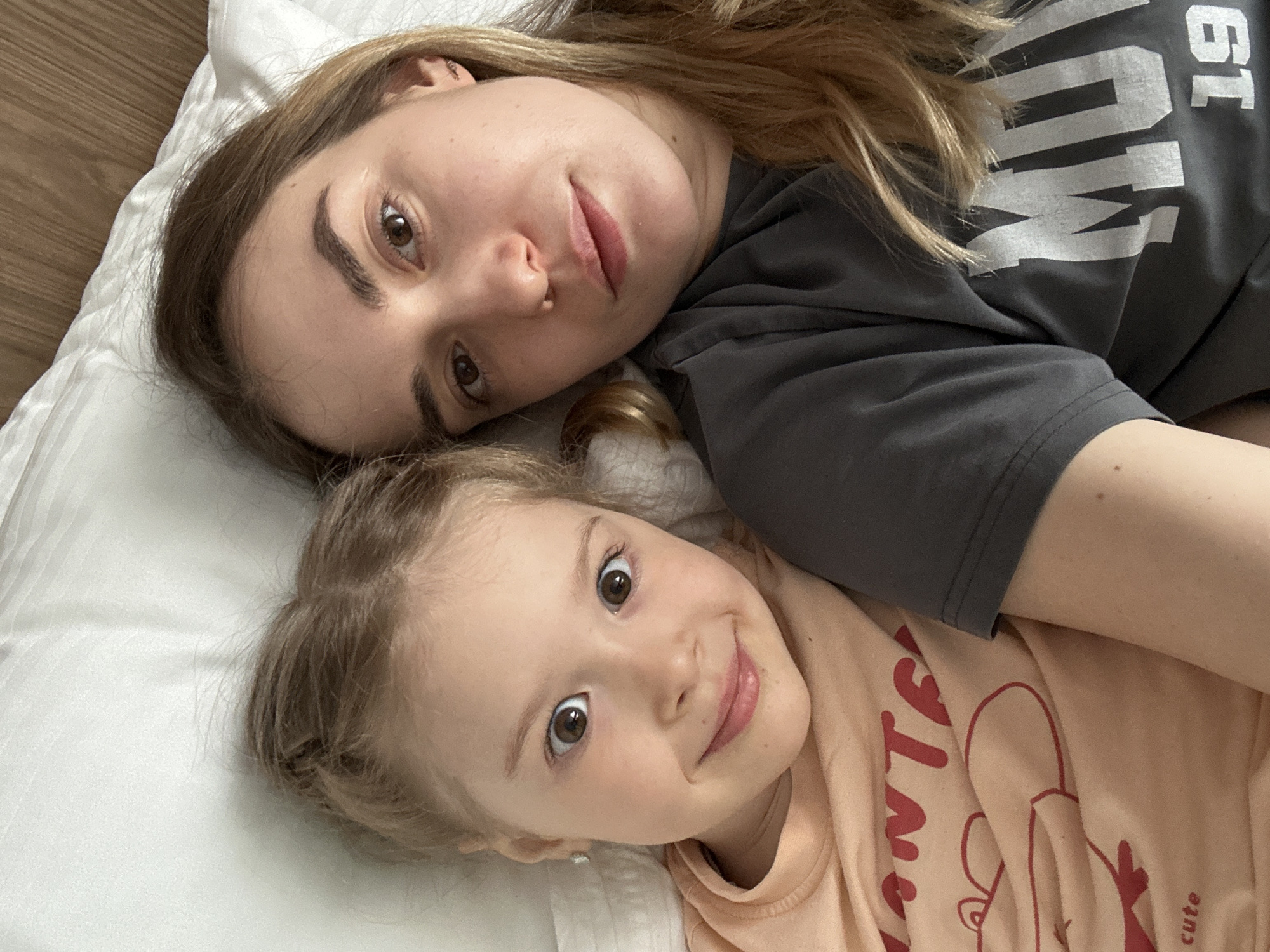 Two individuals, presumably a mother and daughter, are lying on a white pillow or bedding. The woman has brown hair, fair skin, and is wearing a grey t-shirt with white lettering, partially visible as "MU 19." The child has blonde hair, fair skin, and is wearing a light orange t-shirt with a cartoon print. Both are looking at the camera. The environment appears to be indoors, with a wooden floor partially visible in the upper left corner. There is no immediate indication in the image that it was taken in Glazov, Russia.