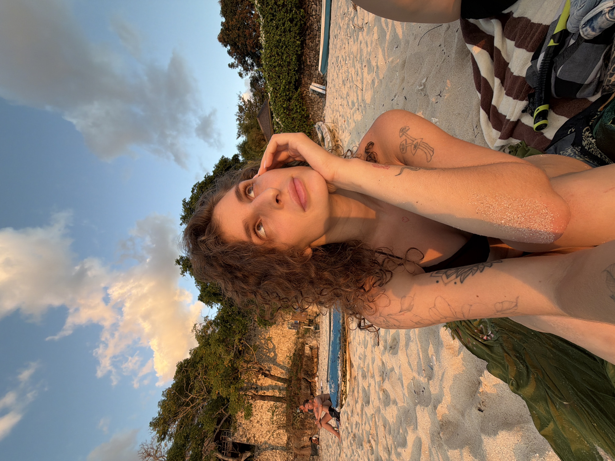A woman with curly brown hair is sitting on a sandy beach, looking up and to the right, with her right hand supporting her chin. She is wearing a black bikini top and has tattoos on her arms and shoulder. In the background, there are trees, a stone wall, and a body of water. To her left, another person is sitting on the sand next to a blue object, possibly a boat or a mat. The sky is blue with scattered clouds, illuminated by golden hour light.