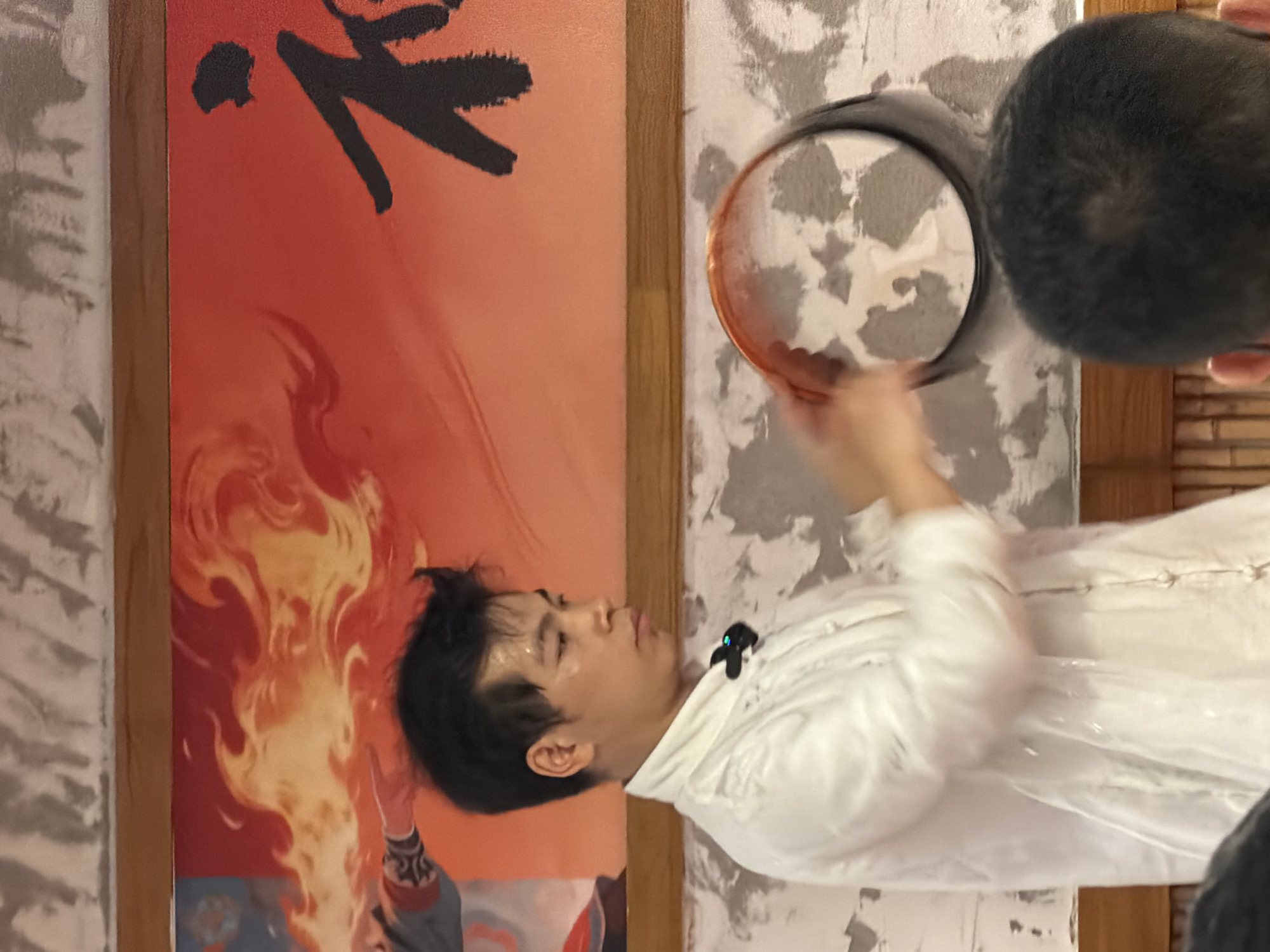In Yuzhong District, China, a young man in a white traditional Chinese garment is captured mid-action, holding a circular object. The man has dark hair and appears to be sweating, suggesting physical exertion. He is looking towards his left. Behind him, a large mural adorns the wall, featuring fiery imagery and what appears to be Chinese calligraphy in black. The wall itself has a textured, light-colored finish. In the foreground, the back of another person's head is visible. The overall setting suggests an indoor space, possibly a cultural performance venue or practice area.
