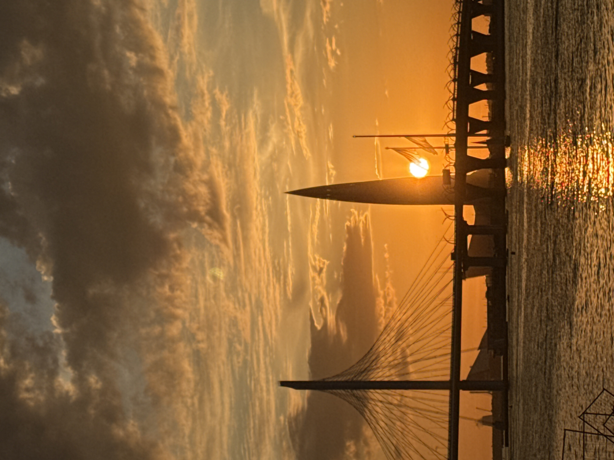 This image captures a vibrant sunset or sunrise over a body of water in Saint Petersburg, Russia. The sky is dominated by a dramatic array of clouds, painted in warm hues of bright orange, yellow, and darker, muted browns, transitioning from the horizon upwards.
A brilliant, circular sun is positioned low in the sky, partially obscured by the silhouette of a large vessel in the middle ground. This vessel, possibly a yacht or a passenger ship, features a prominent mast or superstructure directly in line with the sun, and what appear to be flags are visible near the sun's disc.
Two distinct bridges span the waterway, both silhouetted against the glowing sky. To the left, a modern cable-stayed bridge stands with its tall pylon and numerous fanning cables. To the right, another bridge structure, possibly older with multiple supports or arches, stretches across the water.
The water in the foreground and mid-ground reflects the intense sunlight, creating glittering, shimmering streaks of orange and yellow light on its surface. The scene evokes a tranquil and majestic atmosphere, highlighting the interplay of natural light with man-made structures. No text is visible in the image.
