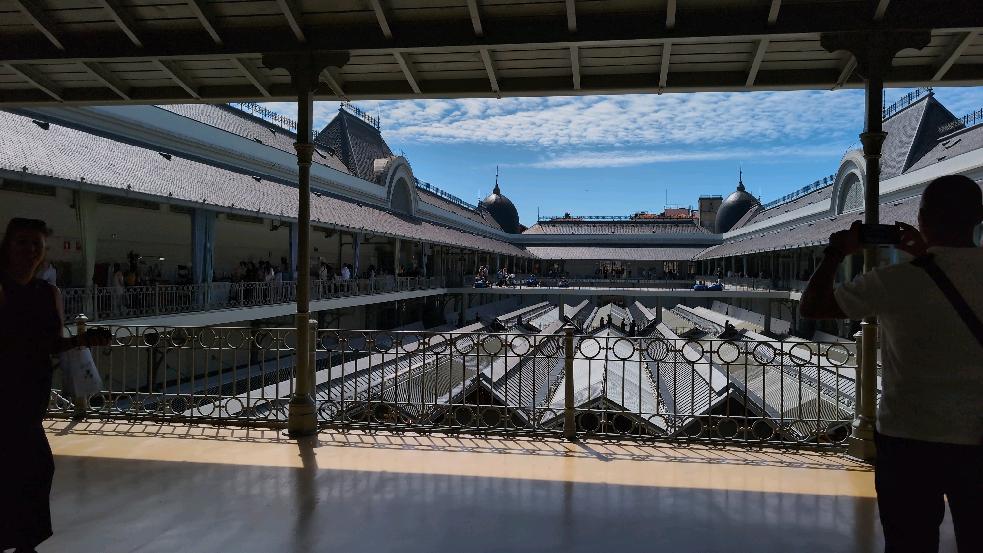 The image shows a view from an elevated walkway overlooking the interior of the Mercado do Bolhão in Porto, Portugal. The market, with its distinctive glass and metal roof structure, is visible below, featuring rows of stalls. People are walking along the upper level walkways and can be seen in the open-air courtyard. The sky is blue with scattered white clouds. In the foreground, on the right, a person is silhouetted against the light, holding up a phone to take a photograph. On the left, a woman is also in silhouette, walking away from the camera. The architecture of the market is grand, with arched windows and ornate railings. The overall impression is a bustling and well-lit indoor market on a sunny day.