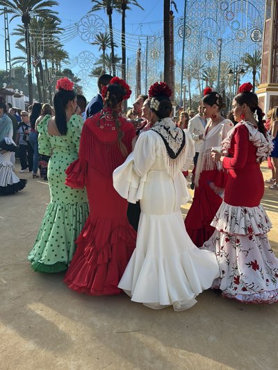 In Jerez de la Frontera, Spain, a group of women dressed in traditional flamenco dresses stands together outdoors. The woman on the left wears a green dress with black polka dots. Next to her is a woman in a red dress adorned with fringe and embroidered floral designs. The third woman is in a white dress with a black and white shawl. The fourth woman wears a red dress with a white floral-patterned flounce. All the women have flowers in their hair. Other people, along with trees and festive string lights, are visible in the background.