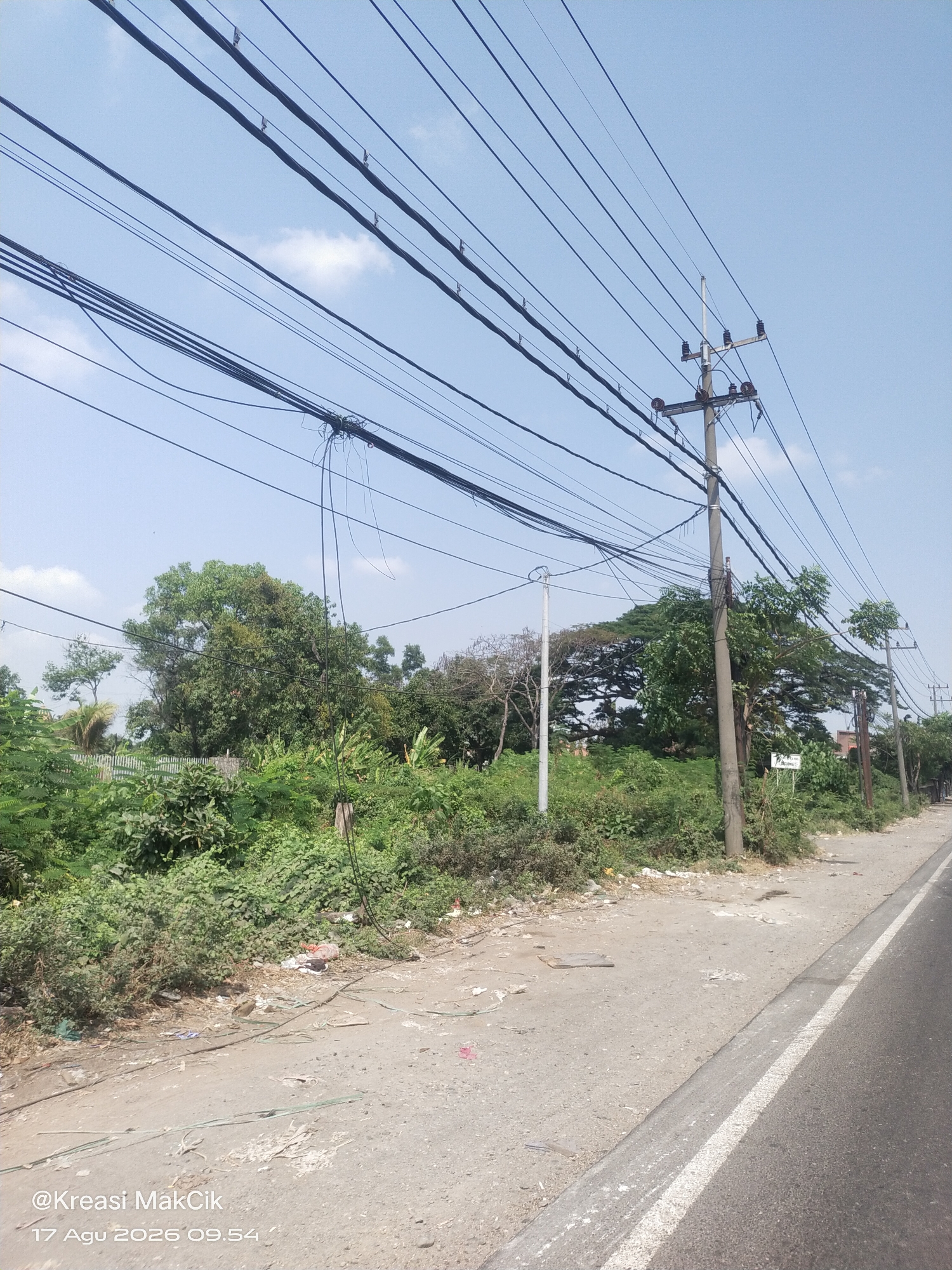 The image depicts a roadside scene in Kletek, Indonesia, with a clear blue sky overhead and a series of overhead power lines dominating the upper portion. A concrete utility pole with multiple insulators and hanging wires stands prominently on the right side. The ground beside the road is a mixture of dirt and gravel, with some scattered debris including plastic bags and papers. Lush green vegetation, including trees and bushes, lines the roadside, with a white picket fence visible in the background behind some of the foliage. A paved road with a white line marking its edge is visible on the right. The timestamp "@Kreasi MakCik 17 Agu 2026 09.54" is superimposed on the lower left corner of the image.
