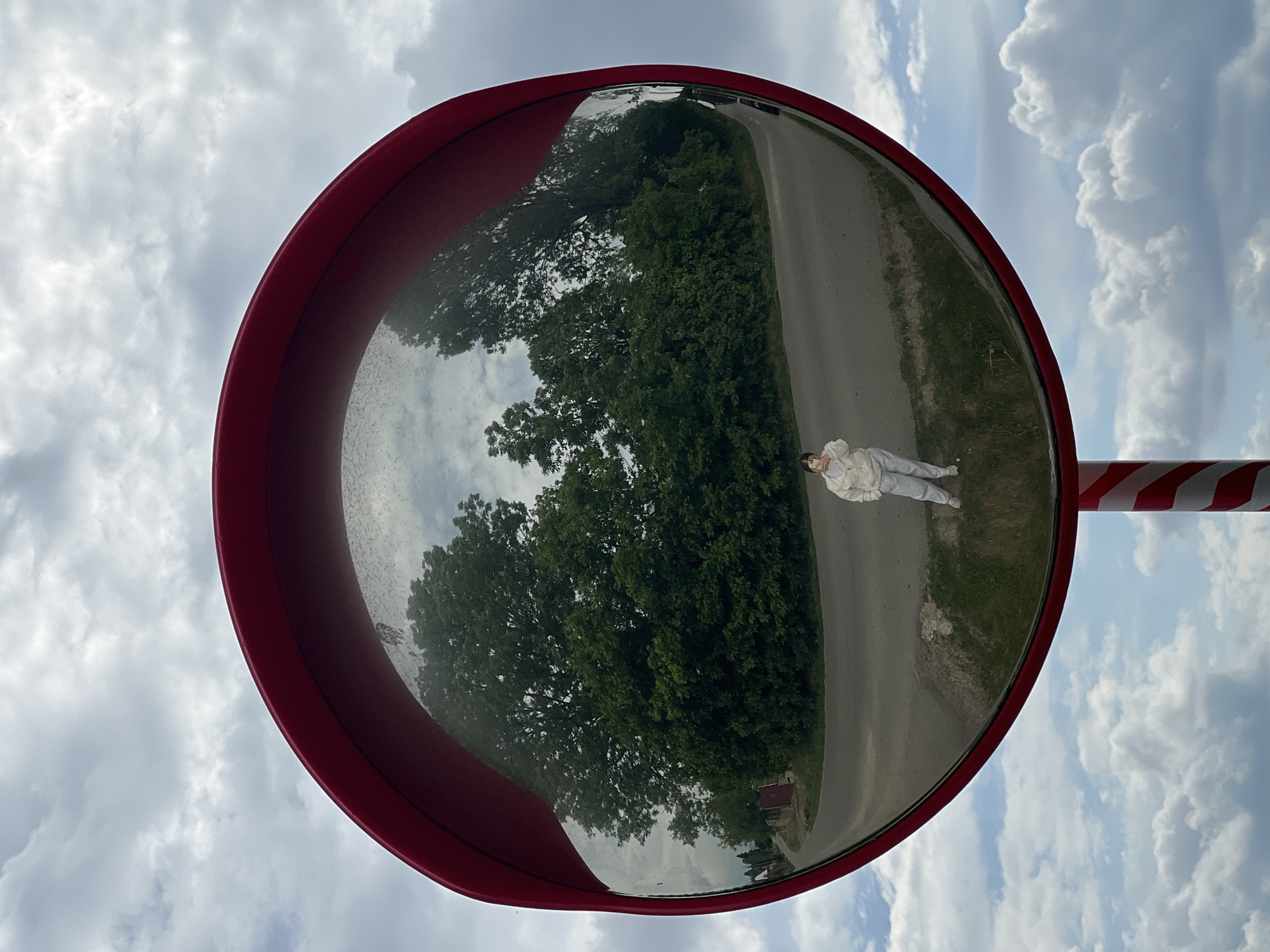 A convex mirror, framed in red, captures a scene in Kozivka, Ukraine. The mirror is mounted on a pole painted with red and white stripes, and it reflects a stretch of road bordered by green vegetation on one side and trees on the other. A person, dressed in light-colored clothing, is visible at the edge of the road within the mirror's reflection. The sky above is partially covered with clouds.