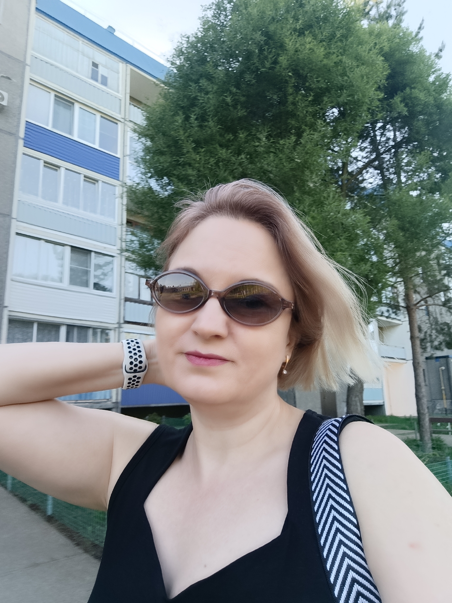 A woman is taking a selfie outdoors. The woman has short blonde hair and is wearing brown sunglasses, a black top, a watch, and a handbag with a white and black striped strap. Her right hand is raised with her wrist near her neck. Behind her is a multi-story apartment building with white and blue panels, windows, and balconies. To the right of the building is a large green tree. The location appears to be in the city of Марковский, Russia.