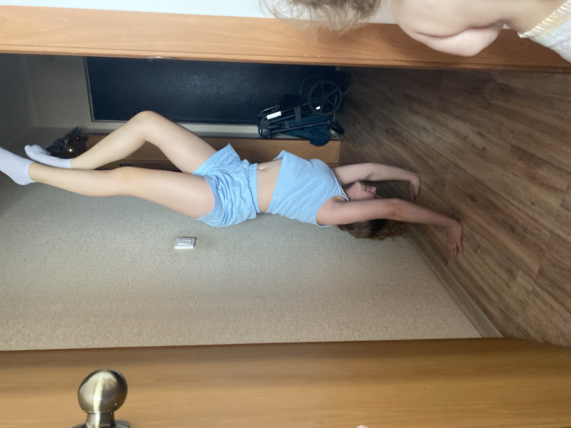 A woman is lying on a carpeted floor, wearing a light blue top and shorts with white socks. She is positioned in a room that appears to be under a loft or an attic space. Her arms are stretched out, almost touching a wall with wood paneling. Above her, there is a dark object, possibly a television or monitor, and a folded stroller. A small ornament is positioned next to her feet. Above and below the scene, there is a blurry depiction of wooden trim, which suggests the image was taken at an unusual angle or through a doorway. The location is unknown.