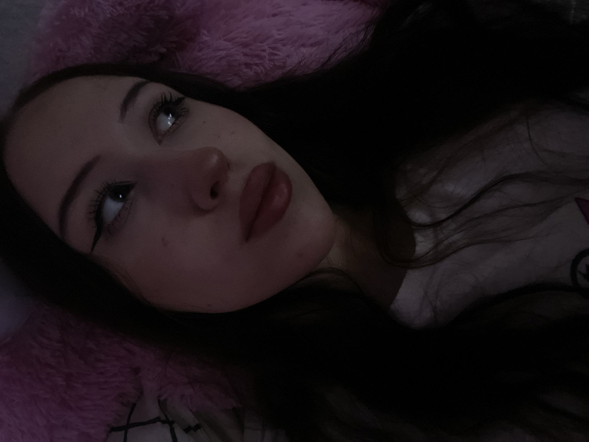 A close-up, overhead shot shows a young woman lying down, with her head resting on a pink, fluffy object. Her dark hair is spread around her. She is looking upwards and to the right, with her eyes open and her mouth slightly parted. Her face is illuminated by a soft light, creating shadows that highlight her features. The background is dark, with only the pink object and a hint of a patterned surface visible. The image does not provide any information about the location in Votkinsk, Russia.