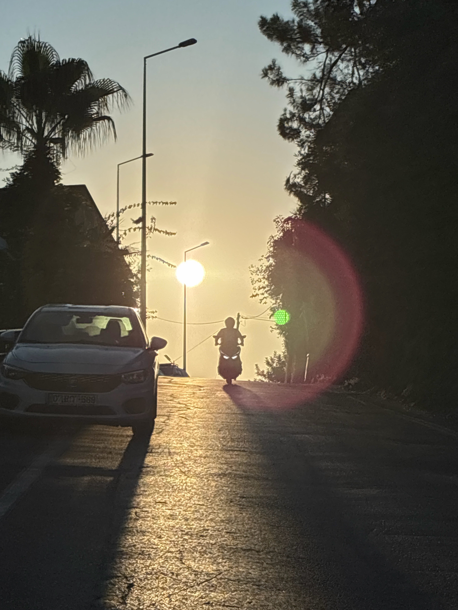 A white car is parked on the left side of an asphalt road that slopes upwards towards a bright sun setting in the distance. The car's headlights are on, illuminating the road ahead and casting a long shadow to its left. A person is riding a scooter up the road, silhouetted against the setting sun. A palm tree is visible to the left of the road, and dense foliage lines the right side. A street lamp is positioned behind the car. The scene is characterized by the warm glow of the sunset, with lens flares creating colorful halos around the light sources. This image appears to have been taken in Alanya, Turkey, based on the presence of Mediterranean flora and the style of the car's license plate which resembles Turkish plates.