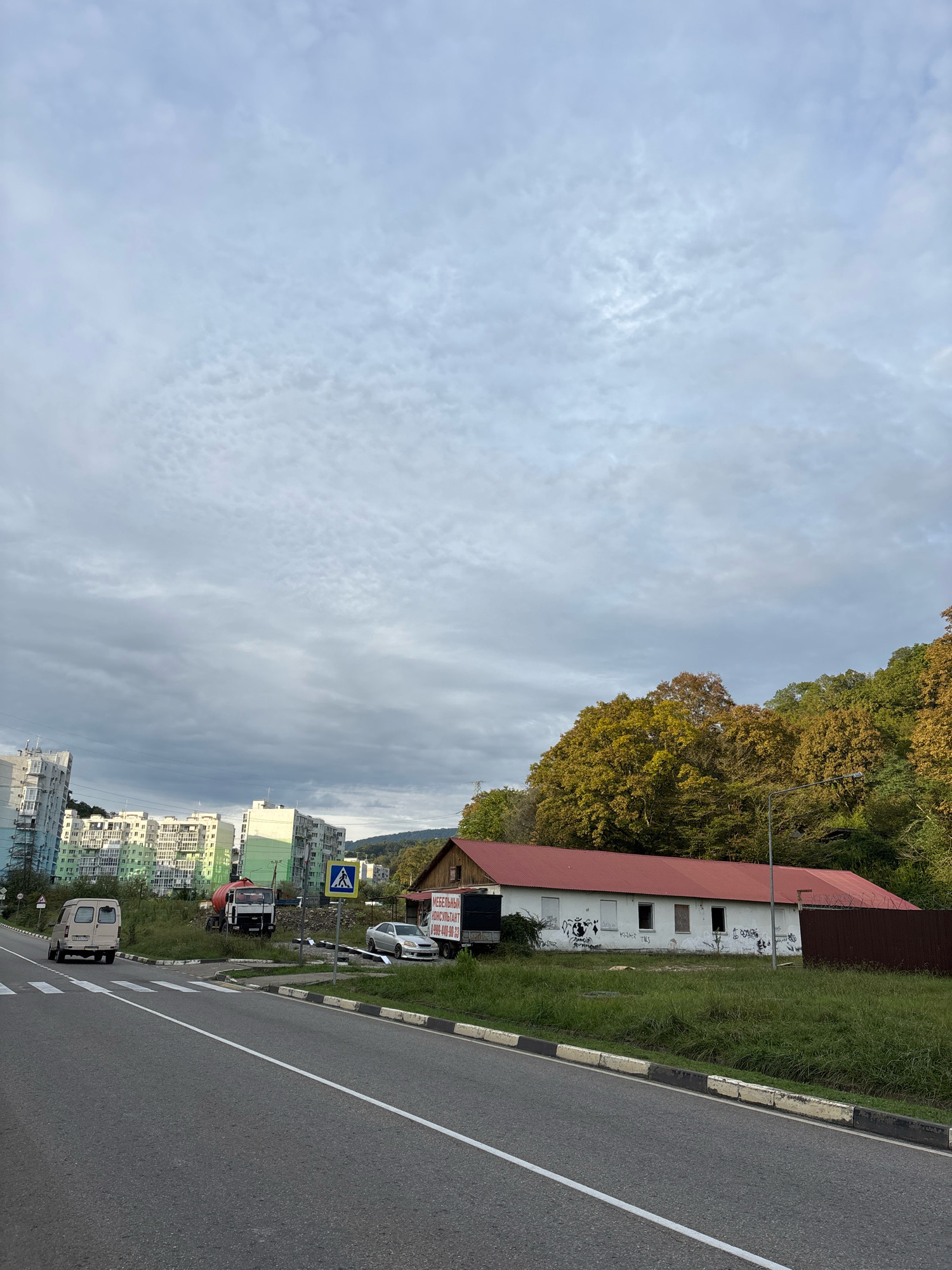 The image depicts a street scene in Sochi, Russia, under a cloudy sky. On the left, a multi-lane asphalt road curves towards the background, marked with white lines and a pedestrian crossing. A white van is parked on the left side of the road. In the middle ground, several multi-story residential buildings with light-colored facades are visible, some with balconies. Adjacent to the buildings, a concrete mixer truck is parked on a grassy area. A pedestrian crossing sign is present near the road.
To the right of the road, a long, single-story building with a red-tiled roof and white walls covered in graffiti stands. In front of this building, a grassy area with some vegetation is present. Behind the building, a densely wooded hillside with trees exhibiting autumn colors can be seen. A brown metal fence runs along the right edge of the property. Further in the distance, a dark-colored truck is parked near the building. The overall impression is of a moderately developed area with a mix of residential and commercial or industrial structures, set against a natural backdrop.