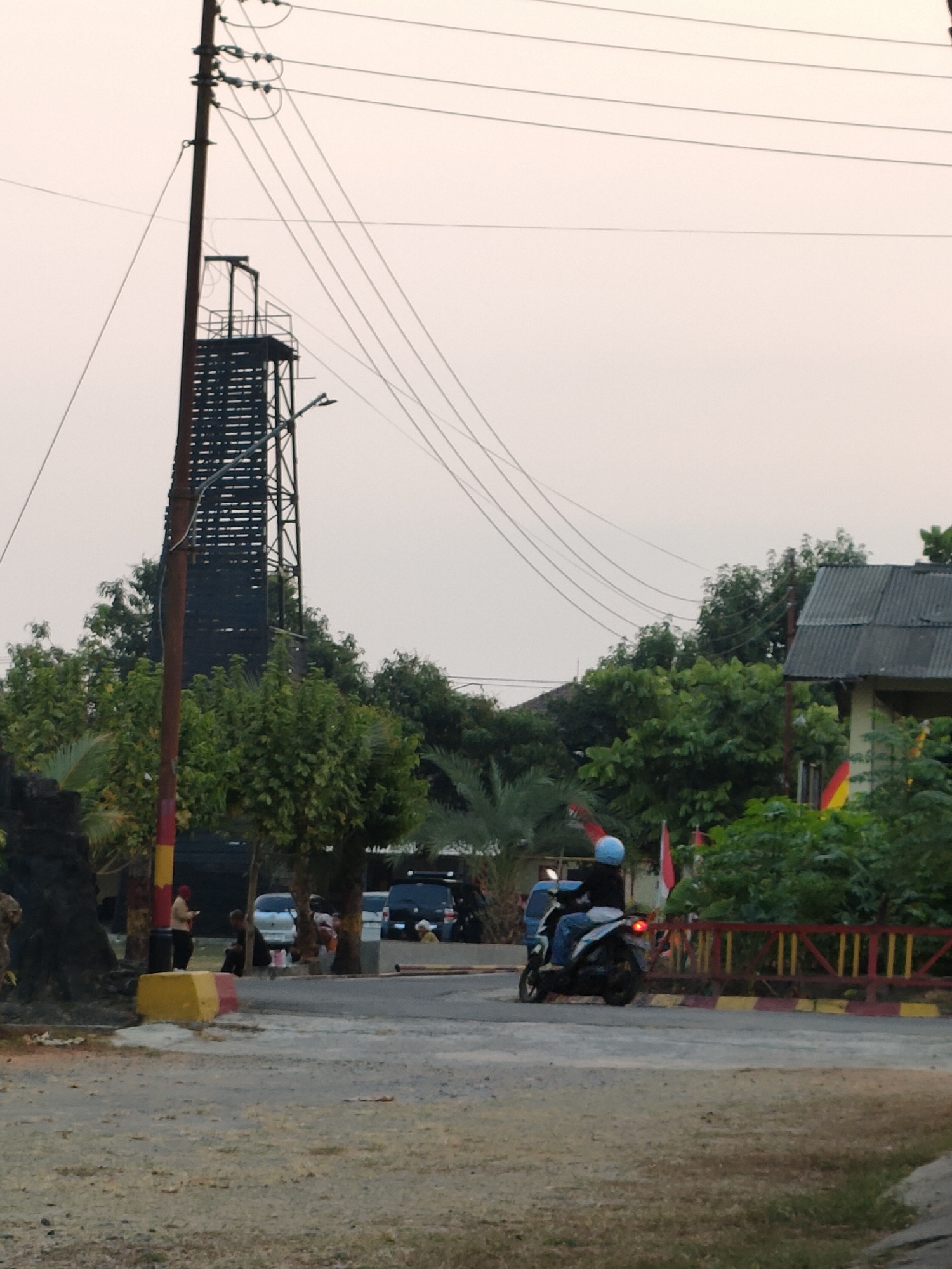 The image depicts a scene on a street in Banguntapan, Indonesia. In the foreground, a dirt and gravel path leads to a paved road. On the right side of the road, a motorcycle with a rider wearing a blue helmet is moving away from the viewer. Behind the motorcycle, a red and yellow striped fence runs along the side of the road, with a building and lush green trees visible beyond it.
Dominating the left side of the image is a tall, dark metal structure resembling a communication tower or observation deck, supported by a reddish-brown pole with yellow and red bands near its base. Several power lines are strung across the sky above the scene.
Further down the road, several vehicles, including a black SUV and a white car, are parked or driving. A few individuals are visible on the sidewalk and near the vehicles, some appearing to be interacting with their phones or talking. The sky is overcast, suggesting either early morning or late afternoon. The overall impression is a moderately busy urban or semi-urban environment.