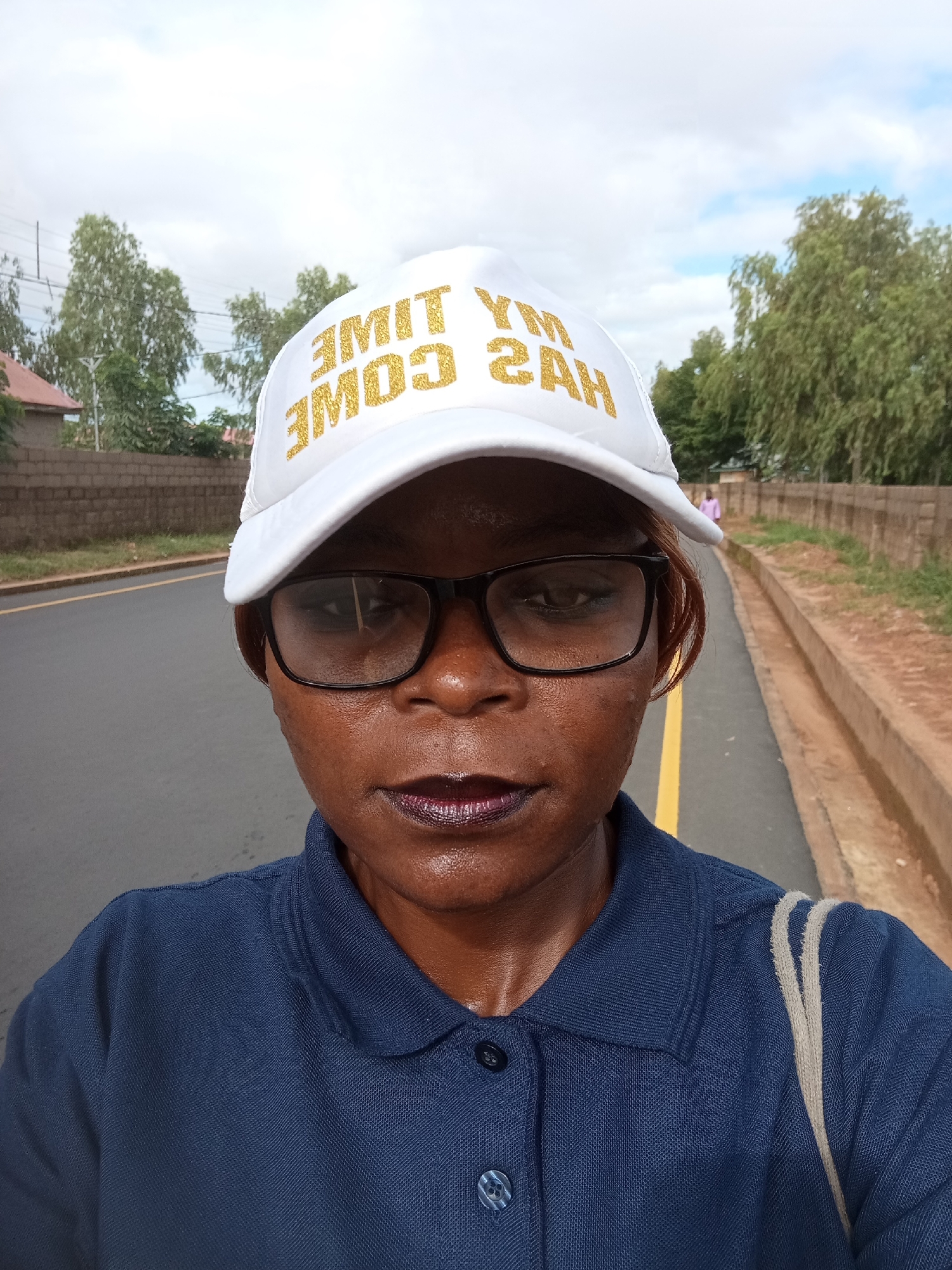 The image depicts a close-up of a Black woman wearing a white baseball cap with gold lettering that reads "MY TIME HAS COME." She is also wearing glasses and a dark blue collared shirt. The background shows a paved road with a yellow line, a brick wall, a drainage ditch, and trees. In the distance, a person in a pink shirt is walking away from the camera. The setting appears to be outdoors, possibly in a semi-urban or rural area. There is no specific information within the image to confirm the location as Mayo Dasa, Nigeria.