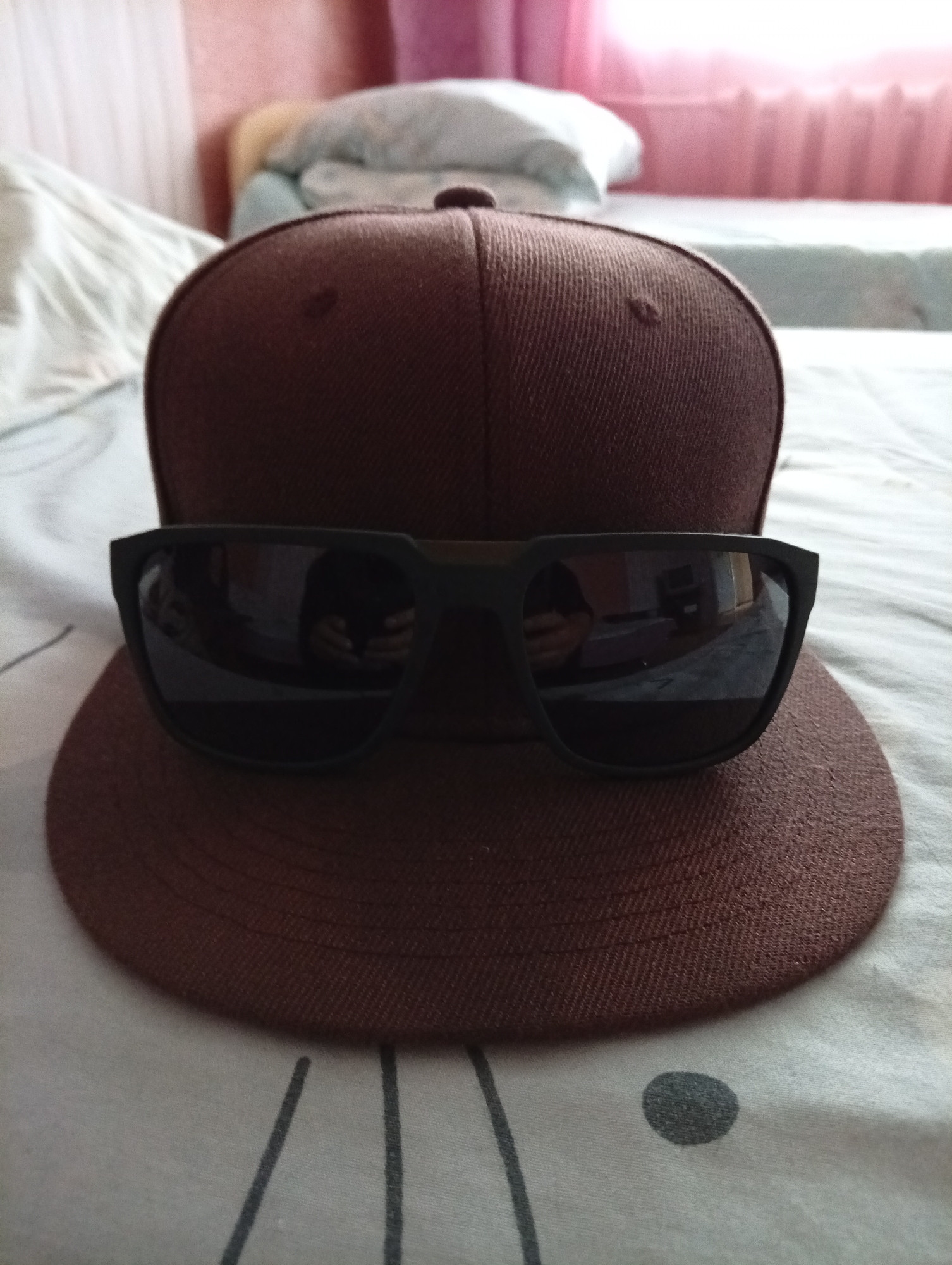 A close-up shot shows a brown baseball cap with sunglasses placed on top of its brim. The cap and sunglasses are positioned on a bed with a white and gray striped comforter. In the background, a pink curtain and a light blue blanket are visible, suggesting an indoor setting, likely a bedroom. There is no indication of Yanaul, Russia, in the image itself.