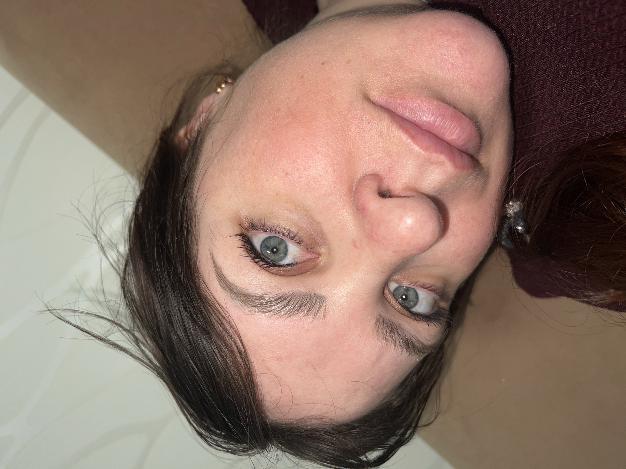 The image presents a close-up portrait of a person, likely an adult female, with the main image oriented upside down. The person has fair skin, light blue-grey eyes, and dark brown hair, which appears somewhat messy and frames the face. Mascara is visible on the eyelashes, and the eyebrows are dark and defined. The lips are full and naturally pink, slightly parted. The tip of the nose shows a hint of redness. A few small moles or freckles are present on the forehead.
The person is looking directly towards the viewer with a neutral to slightly relaxed expression. Two gold-colored earrings with small spherical details are visible in the upper part of the left ear (when viewed upright), and another, possibly reflective, earring is near the right ear.
The background consists of a light-colored, possibly white or off-white, surface on one side, which could be a wall or bedding. On the other side, a dark reddish-brown textured fabric, likely clothing or upholstery, is visible. The lighting is soft and even, suggesting an indoor setting. There are no specific cues to indicate the time of day, nor are there any visible texts or overt location details within the image itself. Based on the provided context, the location is Perm, Russia.