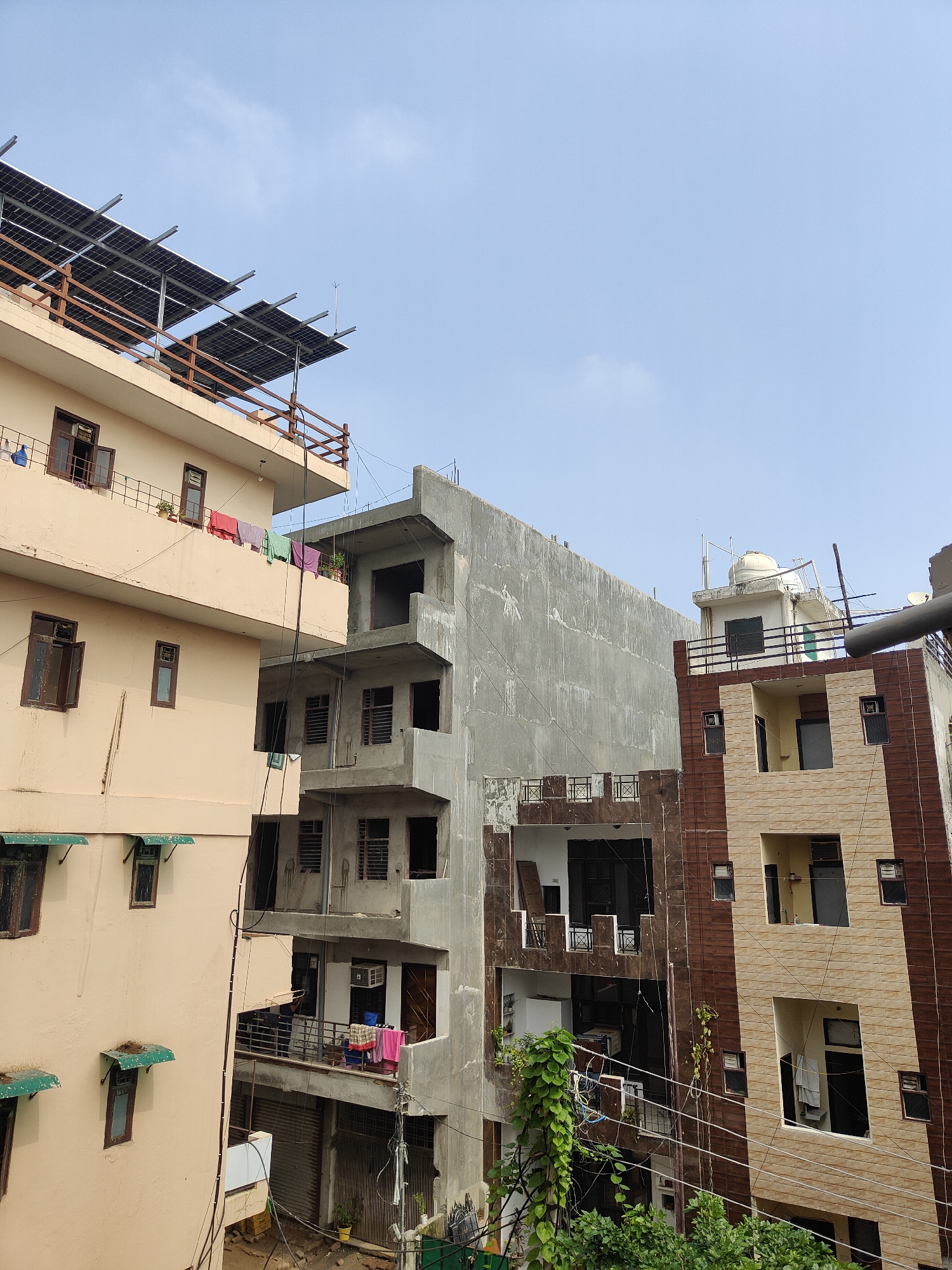 The image shows a collection of apartment buildings in Gurgaon, India. On the left, a light-colored building features solar panels on its rooftop and laundry hanging on a balcony. To its right, a building under construction displays exposed concrete and unfinished balconies. Further to the right, a building with a brown and beige patterned facade has several balconies, some of which are recessed. A water tank is visible on the rooftop of one of the buildings. The sky above is a pale blue with a few wispy clouds.