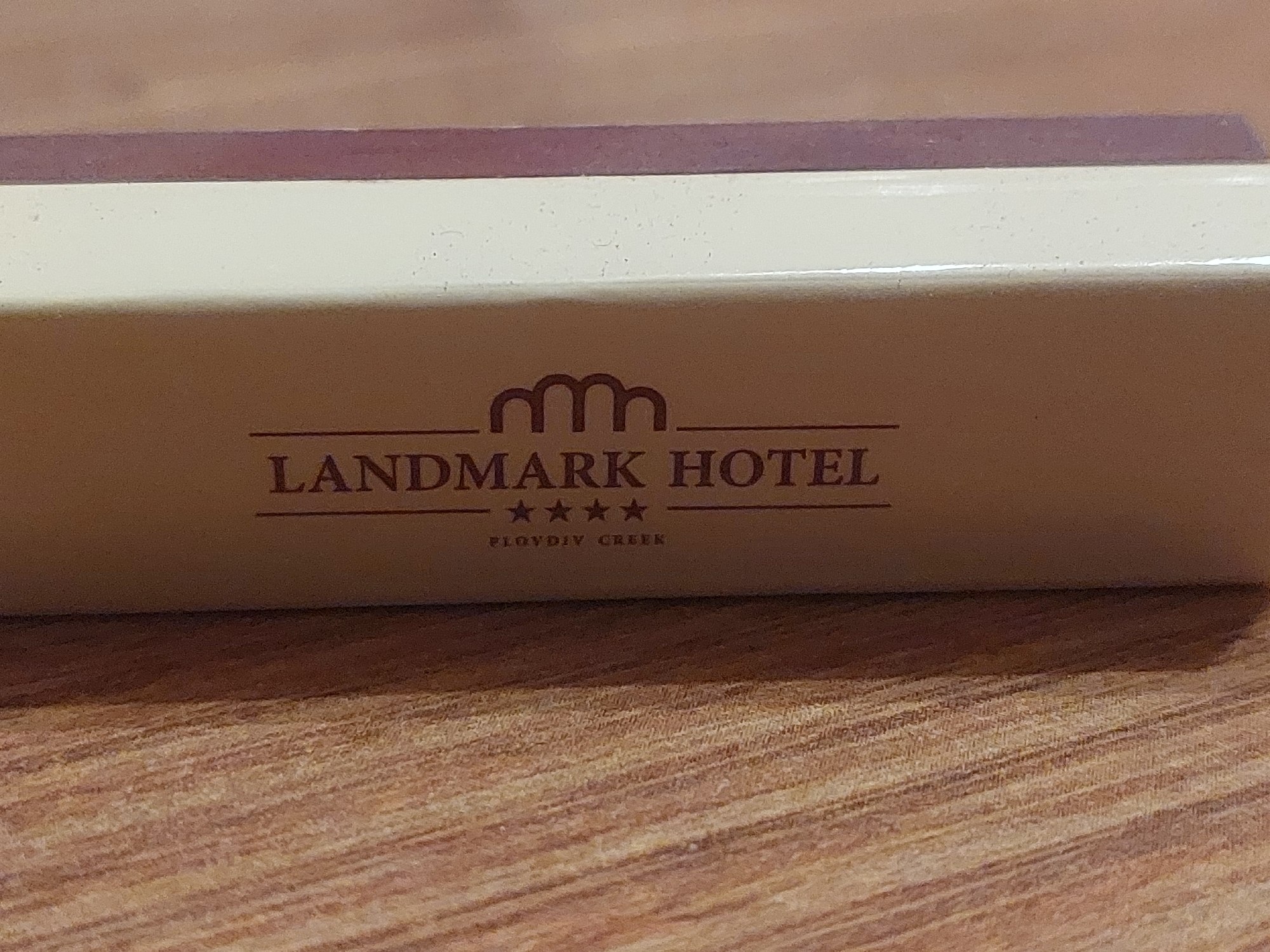 The image shows a close-up of a box labeled "Landmark Hotel" in a brown sans-serif font. Above the hotel name is a logo resembling three interconnected arches. Below the name, four stars are centered, and beneath the stars, the words "Plovdiv Creek" are printed in a smaller font size. The box itself appears to be made of a light beige material with a reddish-brown accent on top. The box rests on a surface with a wood grain pattern. The city is not identified in the image; Plovdiv Creek is referenced. There is no information about the location in Tsarevo, Bulgaria.