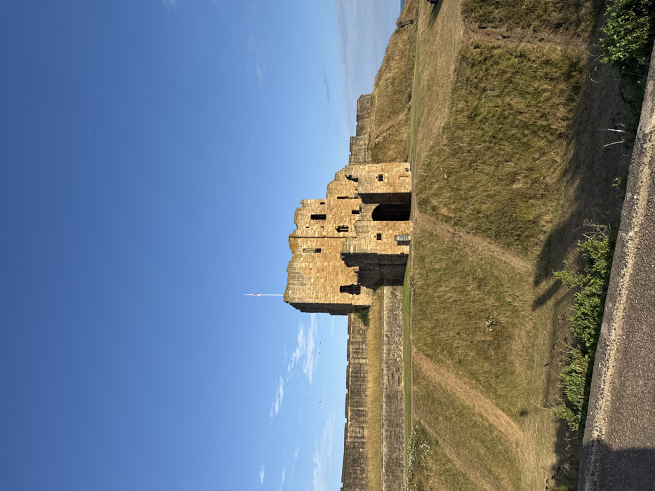 The image depicts a daytime outdoor scene featuring Tynemouth Castle and Priory, located in North Tyneside, United Kingdom.
The primary subject is a large, light brown stone castle structure situated on an elevated grassy embankment, occupying the mid-ground. The castle appears partially ruined, with visible battlements, multiple window openings, and a prominent arched entryway in its lower central section. A white flagpole, topped with a Union Flag, extends from the highest point of the main castle tower on the left. Stone defensive walls extend horizontally to the left from the castle, along the top of the embankment.
The embankment slopes downwards towards the foreground, covered in dry, patchy green and brown grass. Multiple narrow dirt paths are visible worn into the grassy slopes, indicating foot traffic. Long shadows are cast across the lower parts of the embankment and the immediate foreground, originating from the left side of the frame. The immediate foreground shows a grey, textured surface, possibly a paved path or low retaining wall, with sparse green vegetation growing along its edge.
The sky occupies approximately the upper two-thirds of the image, displaying a clear, bright blue hue with minimal cloud cover, predominantly on the right horizon. Several small, dark specks, consistent with birds, are visible in the sky to the left of the castle. A narrow strip of dark blue water, indicative of the sea, is visible along the far right horizon, beyond the castle embankment. A single indistinct, dark figure, possibly a person, stands on the upper grassy area to the far right, near the edge of the frame. A dark sign with white text is affixed to the wall near the castle's arched entrance.