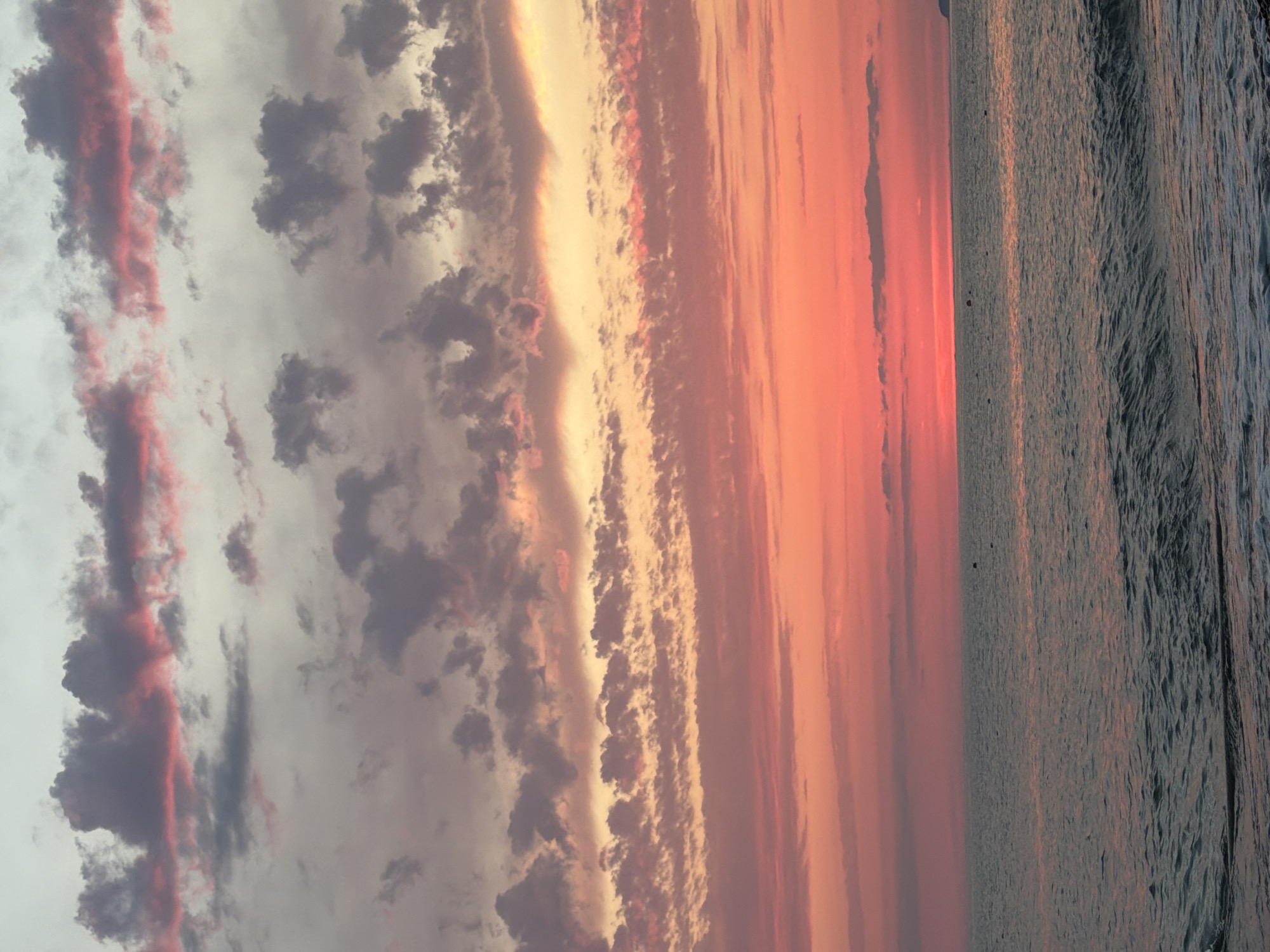 The image depicts a serene seascape at sunset. The sky is a canvas of vibrant colors, transitioning from a soft grey-blue at the top to fiery oranges and pinks near the horizon. Wispy clouds, tinged with pink and gray, are scattered across the sky, creating a dramatic and beautiful effect. The sea is calm, with gentle waves lapping at the shore. The water reflects the colors of the sky, adding to the overall tranquil ambiance. A few buoys are visible in the distance, suggesting the presence of a shipping lane or fishing area. The coastline is not clearly visible, but a faint outline of land can be seen on the far right of the horizon, hinting at a distant shore. The image captures a moment of natural beauty and peace, ideal for contemplation and relaxation.