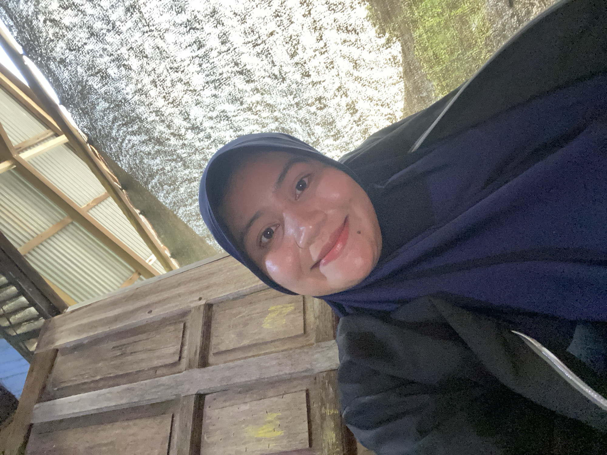 In a low-angle, wide shot, a young woman wearing a dark blue hijab and a black jacket is taking a selfie. Her face, illuminated by natural light, is turned towards the camera, and she is smiling. Her skin appears dewy, and she has a mole on her chin. Behind her, on the left, is a weathered wooden wall with panels. To the right, a corrugated metal roof is visible, supported by wooden beams. A textured black mesh screen covers the area to the right of the woman, allowing sunlight to filter through, creating a dappled effect. The environment suggests an outdoor setting, possibly a rustic building or shelter. This scene is located in Banjarmasin, Indonesia.