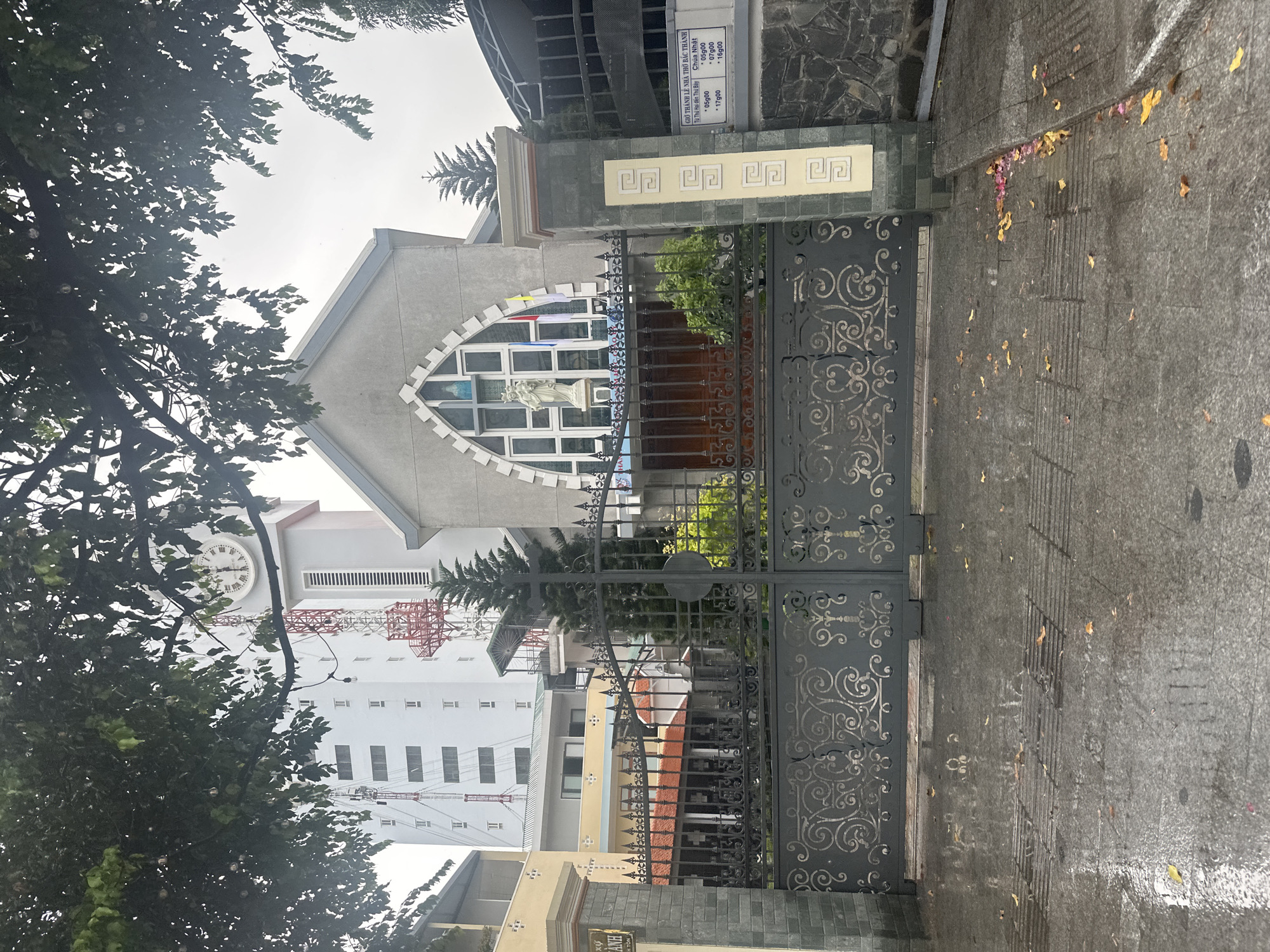 The image depicts a church located in Nha Trang Ward, Vietnam. The church is a grey building with a distinctive, diamond-shaped window featuring a statue. An ornate, wrought-iron gate stands in front of the church entrance. A sign attached to the structure displays religious information and service times in Vietnamese. In the background, taller buildings and a construction crane can be seen. The foreground shows a paved area or sidewalk with fallen leaves. A portion of a tree is visible on the left side of the frame.