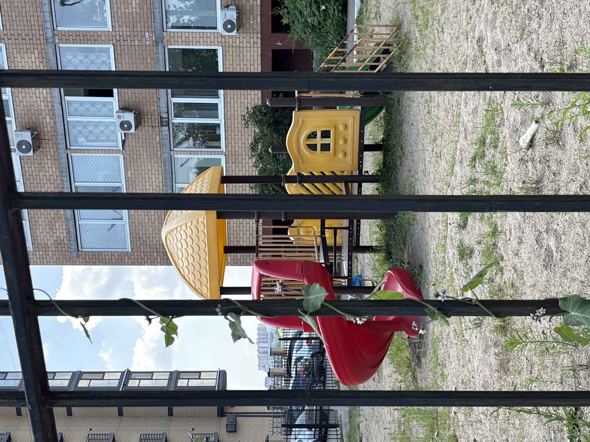 The image depicts a children's playground situated in the city of Blagoveshchensk, Russia, as observed through a black metal fence. The playground features a red spiral slide, a yellow play structure with a roof, and a yellow panel resembling a small house with a window. Green vegetation, including a climbing vine with small white flowers, is growing around the fence and the playground equipment. In the background, a multi-story brick building with air conditioning units and windows is visible, along with several parked cars behind the fence and a distant cityscape. The ground of the playground is covered in sand and sparse grass.