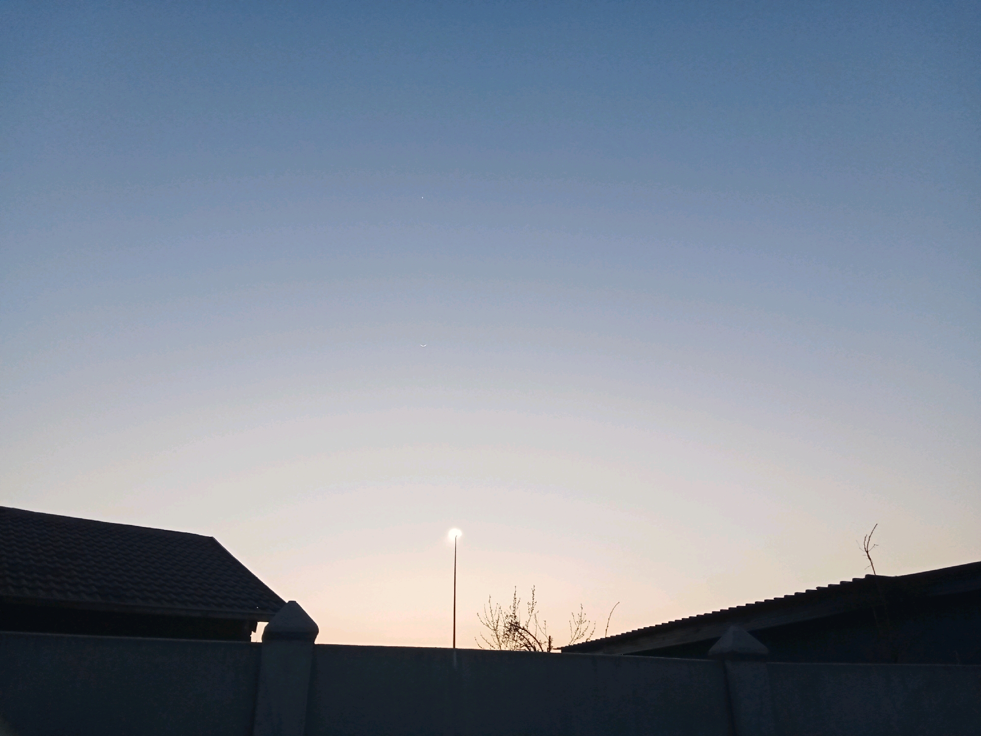 The image shows a suburban skyline at dusk in Meloding, South Africa. Silhouetted against the pale sky are the roofs of houses, a wall, and a street lamp. A faint crescent moon and a distant star are visible in the upper part of the sky. The sun appears to be just below the horizon, casting a soft glow. The overall impression is one of a quiet evening in a residential area.