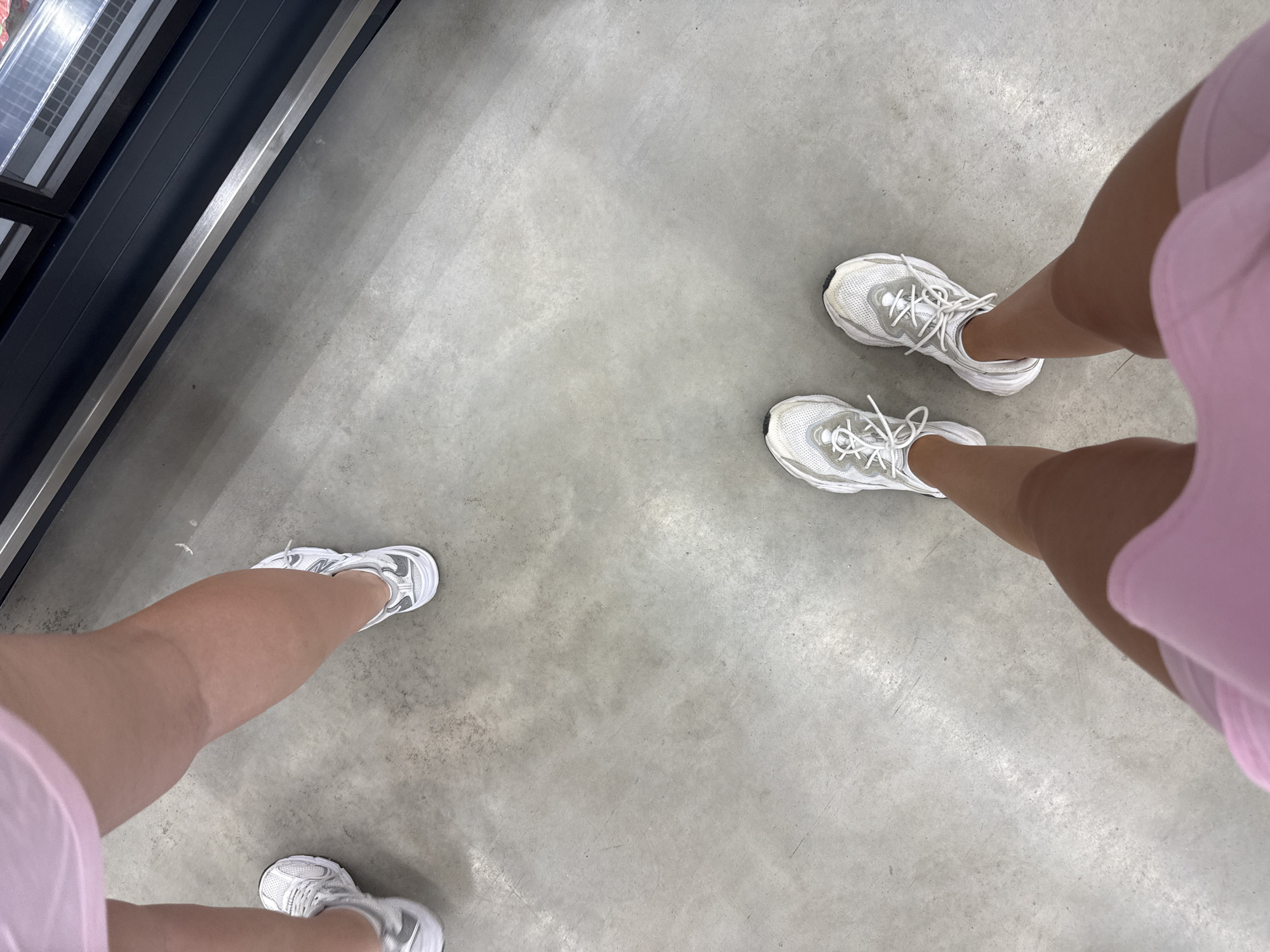 Two individuals are standing on a concrete floor, facing a commercial display case. One person, on the left, is wearing pink shorts and white sneakers. The other person, on the right, is wearing a pink top and white sneakers. The display case appears to be refrigerated, with glass doors showing products inside. The background suggests an indoor commercial setting, possibly a grocery store or a retail establishment. The lighting is bright, casting minimal shadows on the concrete floor. There are no specific landmarks or architectural details that would help pinpoint the exact location within Basarabeasca, Moldova.