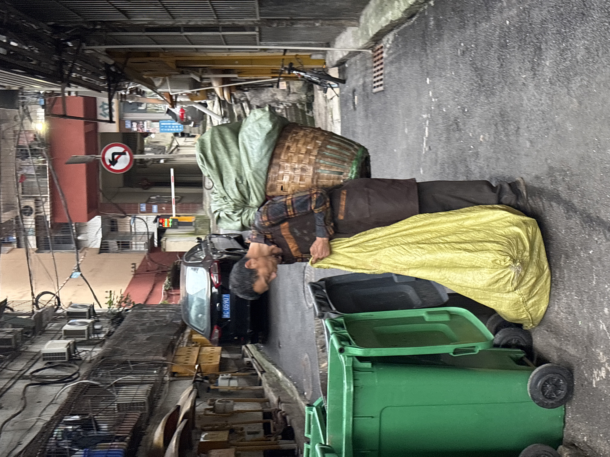 In the city of Jiangjin, China, a man with dark hair and weathered skin is engaged in the task of collecting or disposing of refuse. He is wearing a plaid long-sleeved shirt in brown and orange tones, a dark brown apron or vest, and dark trousers. A large, woven basket is strapped to his back, filled with green material, possibly fabric or plastic sheeting, and covered with more green material. He is holding a bright yellow plastic bag, which appears to be full, with his right hand. His attention is directed downwards towards the bag. He stands beside a row of large, green plastic trash receptacles, one of which has an open lid. Behind him, a black sedan with a license plate reading "Q C 05907" is parked. The scene unfolds on a narrow, paved street flanked by multi-story buildings. The architecture suggests an urban residential area, with balconies, visible air conditioning units, and electrical wiring. A traffic sign with a red circle and a black arrow indicating "no left turn" is visible in the background. The ground is wet, suggesting recent rain. The overall impression is one of a back alley or service lane within a densely populated urban environment.