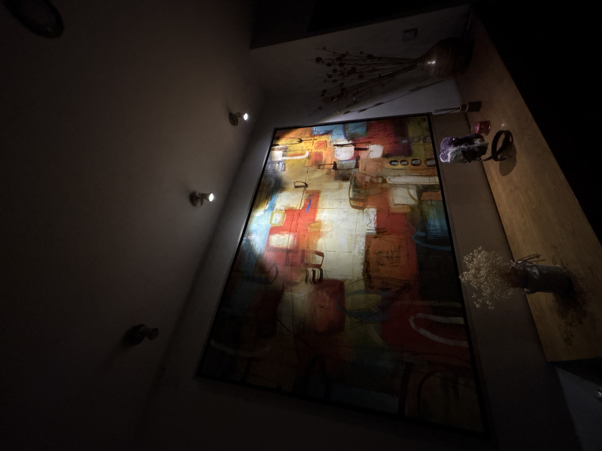 A low-angle shot depicts an interior scene, seemingly a room with artwork and decor. The wall on the left is dimly lit and features multiple recessed lighting fixtures. Dominating the scene is a large, framed abstract painting, rich in colors like red, yellow, and blue. The painting is illuminated, creating a contrast against the dark background.
To the right of the painting, a wooden shelf or table is visible, holding decorative items like a vase with dried flowers and other miscellaneous objects. A decorative vase with twigs or branches extends towards the top of the frame. The room is generally dark, with light sources strategically placed to highlight specific areas and objects.
There's no immediate indication that this image was taken in San Bernardino Tlaxcalancingo, Mexico. It's a straightforward interior shot of a room with art and decor.