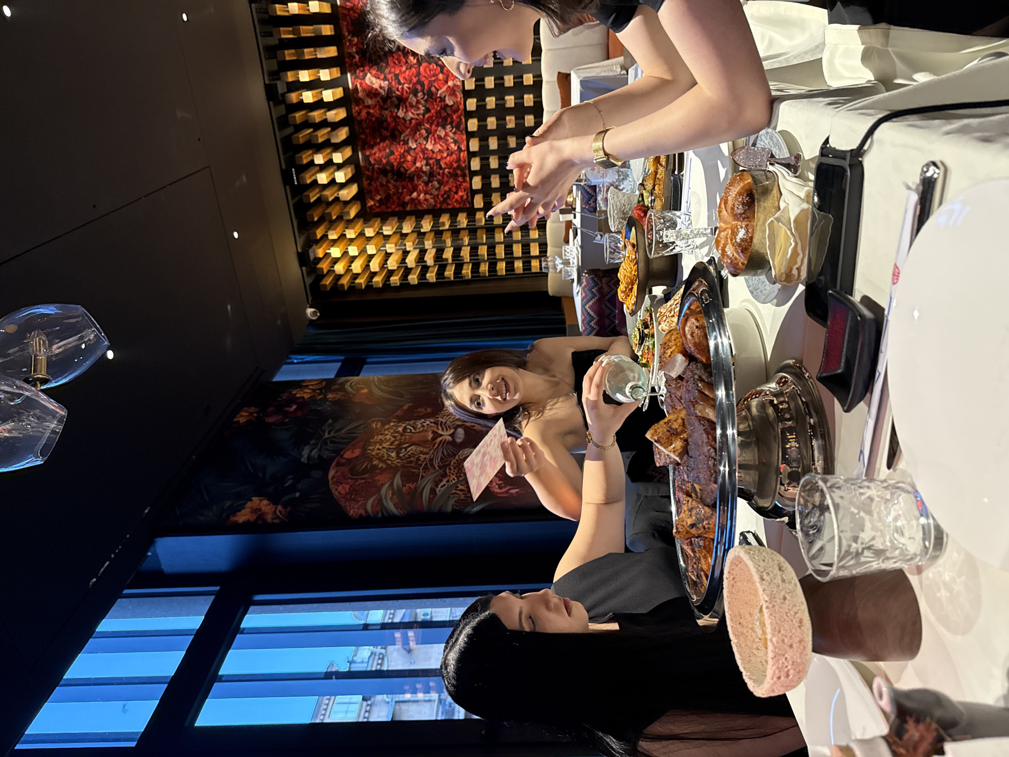 The image shows a dining table in Yerevan, Armenia. Three women are visible around the table. One woman is handling cards, another is poised with her hands above the table, while the third is out of focus. Various dishes of food, drinks, and tableware occupy the table. The background includes a window with a cityscape view, decorative wall panels, and hanging lights.