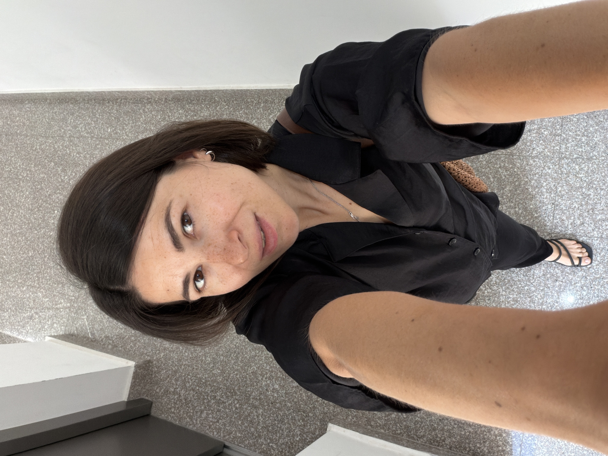 A woman takes a selfie from a high angle looking down. She wears a black button-up shirt and black pants, with black sandals visible on one foot. She has dark hair, fair skin, and freckles. A silver necklace is visible around her neck, and she wears small hoop earrings. The background appears to be a tiled floor and a staircase with white risers and a gray tread. The location is not identifiable as the city of Budva, Montenegro from the image alone.