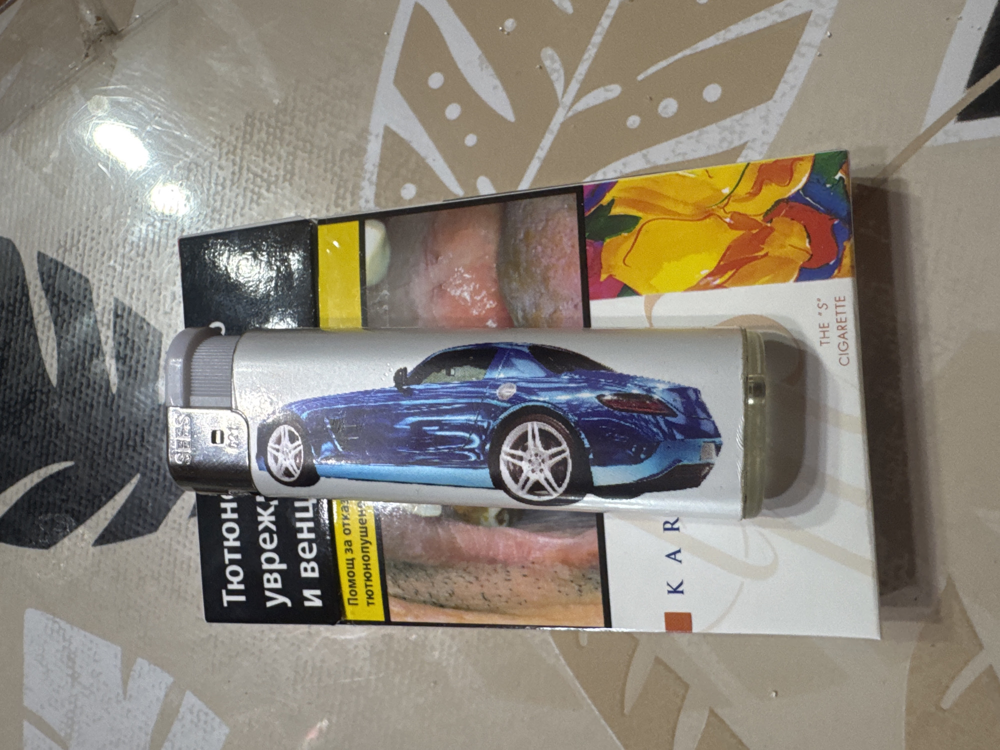 The scene is located in the city of Sarnevo, Bulgaria. An overhead shot depicts a white cigarette pack and a silver lighter with a blue car design, resting on a beige and grey patterned table surface.
The cigarette pack, oriented slightly diagonally from top-left to bottom-right, is predominantly white with a black section at the top containing Cyrillic text that reads 'Тютюн,' 'увреж,' and 'и венц,' indicating tobacco harm to gums. Below this, a yellow band displays 'Помощ за отказ тютюнопушене' (Help to quit smoking). The bottom right of the pack features a vibrant, abstract floral design in yellow, orange, red, blue, and green. Visible text on the lower part of the pack includes 'KAR' on the left and 'THE "S"' 'CIGARETTE' on the right. The pack also displays graphic health warnings: one showing diseased teeth and gums, and another depicting a severe oral or throat lesion.
A silver disposable lighter stands upright on the cigarette pack, obscuring its central portion. The lighter's body is adorned with a graphic of a blue sports car, viewed from above, featuring prominent silver multi-spoke wheels. The top of the lighter has a grey plastic thumb lever and a metal striking mechanism. 'SHES' (or similar text) is embossed on the metal top, along with a vertical mark containing '021' or similar numbering.
The underlying surface is a glossy, textured material, possibly a laminated tabletop or tablecloth, in shades of beige and light grey, patterned with abstract white and darker grey/black floral or leaf-like designs and scattered white dots. Bright reflections on the lighter, cigarette pack, and table surface suggest an artificial overhead light source. No individuals or active interactions are present in the image.