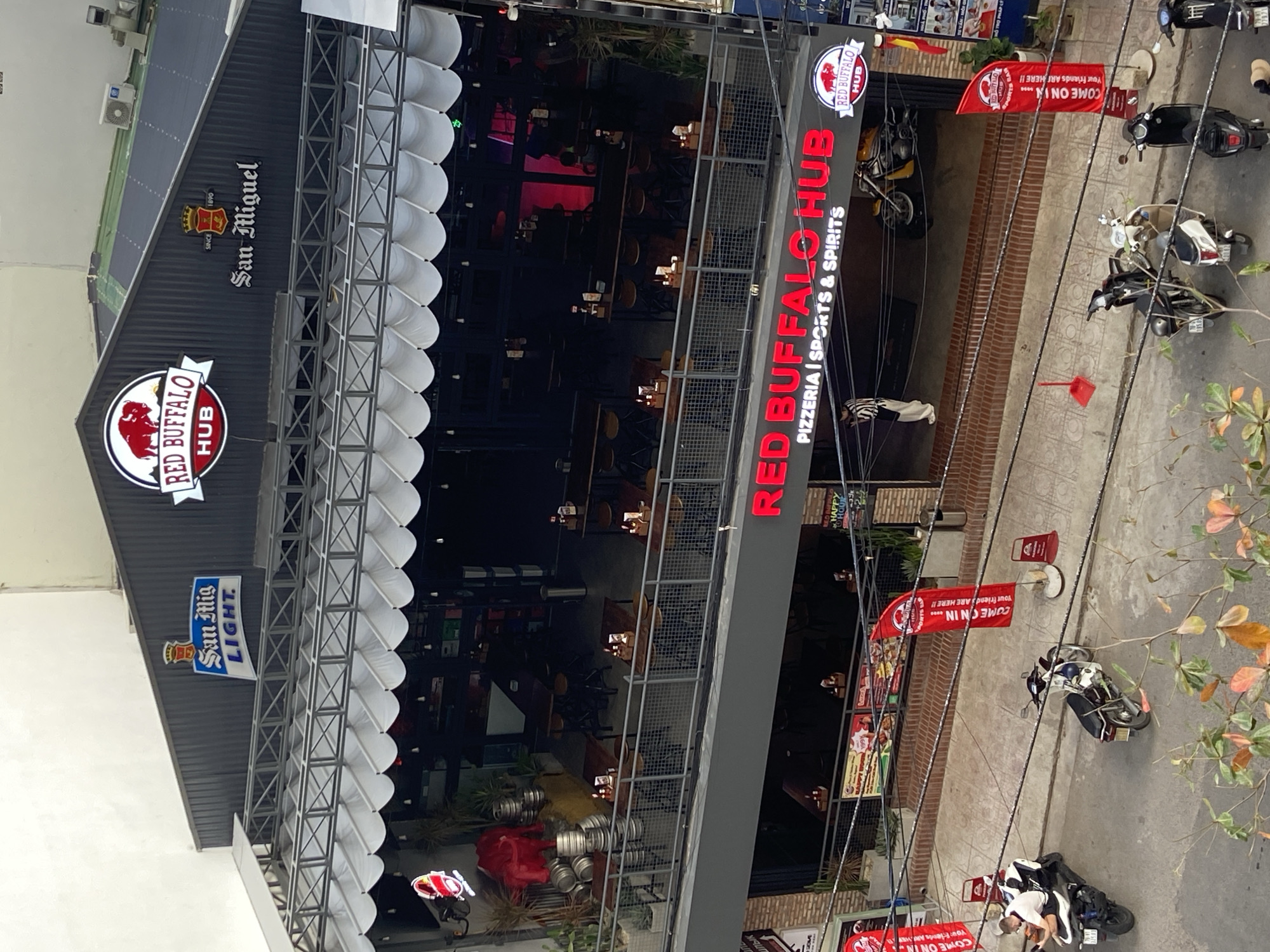 The image shows an outdoor view of a multi-level establishment named "RED BUFFALO HUB" in Bac Nha Trang Ward, Vietnam. The building has a dark-colored corrugated metal facade on the upper levels, adorned with prominent signage. On the top level, the "RED BUFFALO HUB" logo is visible, along with logos for "San Miguel" and "San Mig Light." A white awning with scalloped edges extends over the second level, which features outdoor seating with tables and chairs. The ground level also has outdoor seating and is marked by red banners that read "COME ON IN YOUR FRIENDS ARE HERE!!". Several motorcycles are parked on the street in front of the establishment. A person in a black and white striped shirt and white pants is standing near the entrance on the ground level. The environment appears to be a city street with a sidewalk.
