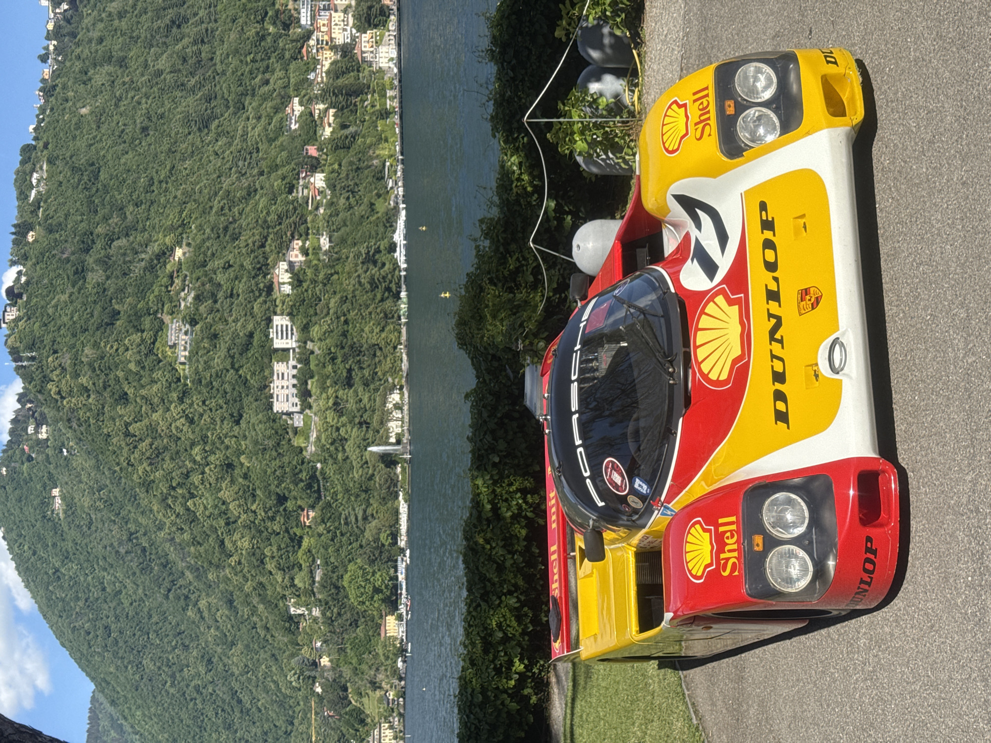 The image showcases a Porsche 962C race car, liveried in the Shell-Dunlop racing colors. It is parked on a paved area, presumably near Lake Como, Italy. The background features Lake Como, surrounded by a densely wooded hillside with scattered buildings. The car's exterior is predominantly yellow, red, and white, with prominent Shell logos and Dunlop lettering. The cockpit is covered and reflects the surrounding scenery. The scene is well-lit, indicating a sunny day. The car appears to be stationary.