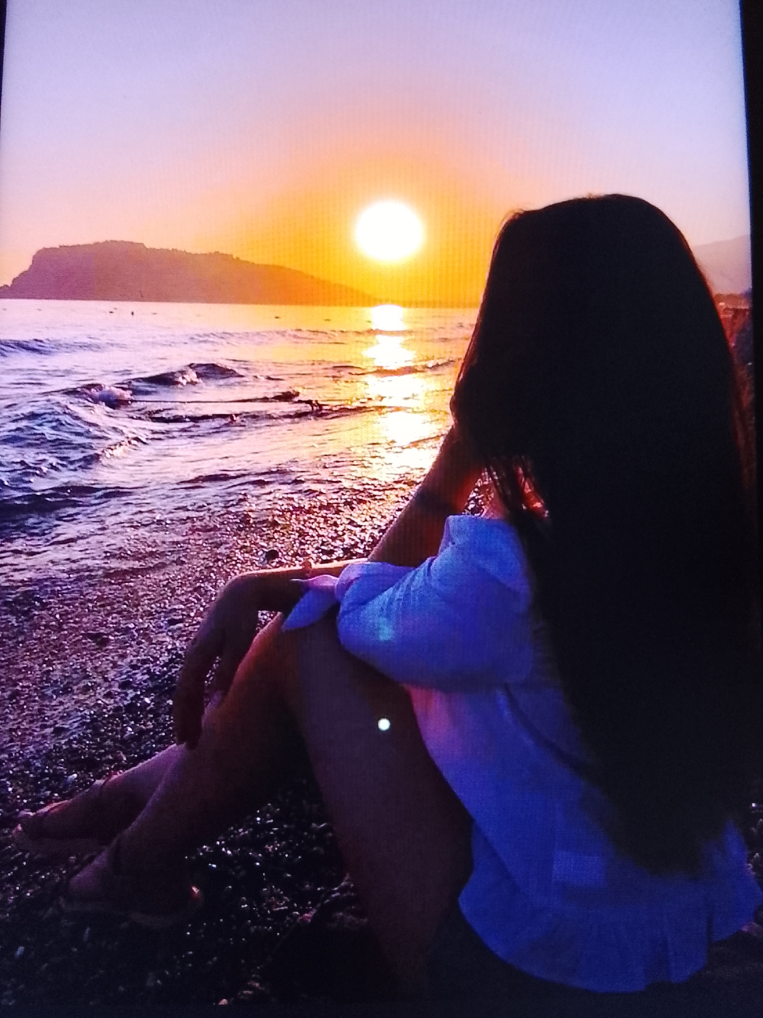 A person with long dark hair is sitting on a pebble beach, facing away from the camera, looking out at the ocean during sunset. The sun is a bright orb on the horizon, casting a warm glow across the water and sky, which transitions from orange to purple. A silhouette of a landmass is visible in the distance. The person is wearing a light-colored shirt. The scene is peaceful and scenic. The provided image does not contain any contextual information that would suggest the location is in the city of Sutki, Russia.