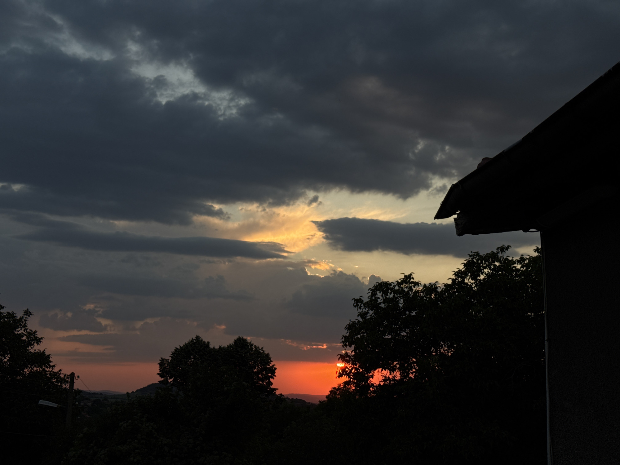 The image captures a dramatic sunset scene viewed from a residential area in Krushevo, Bulgaria. Dark, brooding clouds dominate the upper portion of the frame, with a sliver of vibrant orange and yellow light breaking through near the horizon. In the foreground, the silhouetted shapes of trees and the corner of a dark-colored building with a slanted roof obscure the lower part of the landscape. The building's rough, textured surface is visible on the right side of the image. The sunset casts a reddish-orange glow over the distant hills and the tree line, creating a stark contrast with the dark, stormy sky. The overall impression is one of impending twilight and a powerful, natural display of color.
