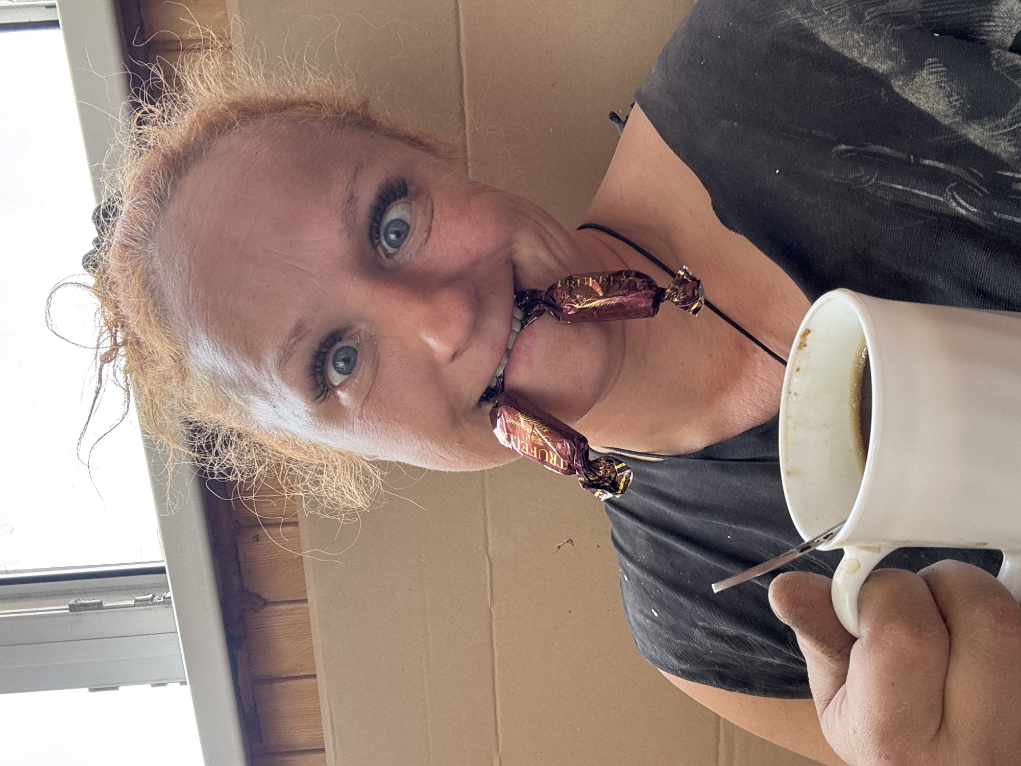 A middle-aged woman with red hair holds a white mug in her right hand while biting down on two wrapped candies suspended from a black cord around her neck. The candies are labeled "TRUFFEL" in Cyrillic script. She has light blue eyes and is wearing a dark-colored shirt. The background consists of a window with a wooden frame and a beige cardboard box. The location is specified as Chaikovsky, Russia, though no element within the image confirms this.