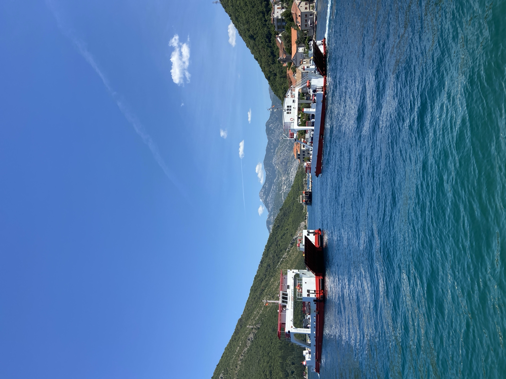 In Lepetane, Montenegro, a clear blue sky stretches above a tranquil, turquoise bay. Two ferries, distinctive with their white upper decks and red hulls, navigate the waters. The ferry on the left is positioned further back, while the one on the right is closer to the viewer, leaving a visible wake. In the background, lush green mountains rise steeply, dotted with terracotta-roofed buildings that cling to the hillsides. Further in the distance, more mountain ranges are visible under the bright sun. The water's surface is rippled, reflecting the vivid colors of the sky and surrounding landscape.