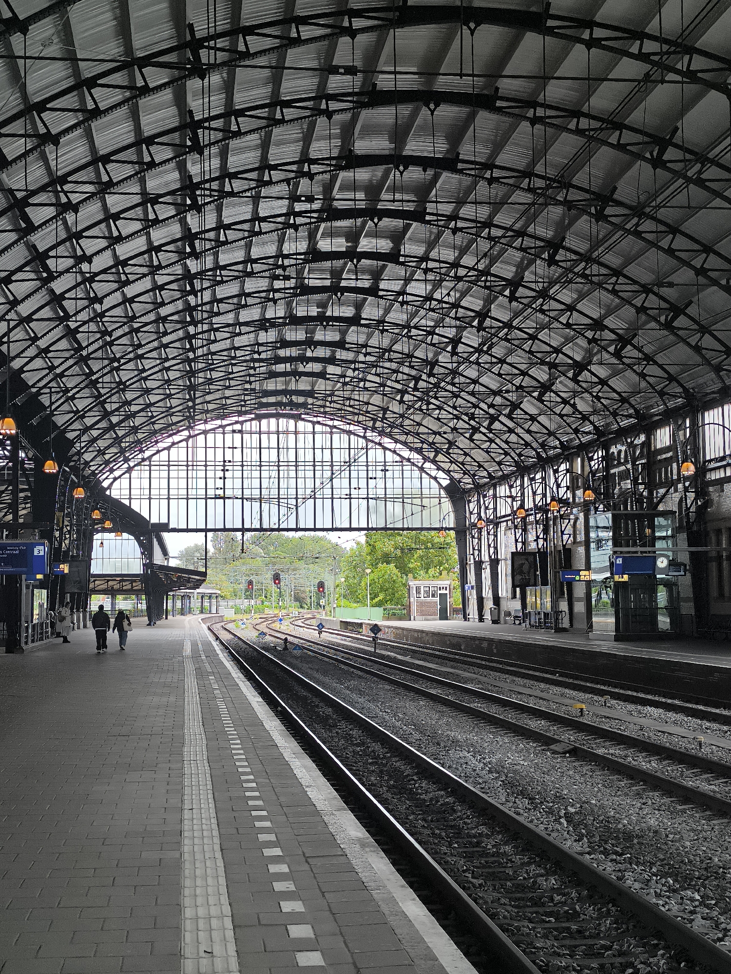 The image depicts the interior of a train station in Haarlem, Netherlands, characterized by its grand, arched metal roof structure. The roof is supported by a complex network of beams and crossbars, with corrugated metal sheeting forming the canopy. Numerous orange-tinted lamps are suspended from the roof. A large, arched glass window dominates the far end of the station, offering a view of greenery and trees outside.
In the foreground, a tiled platform runs alongside multiple sets of train tracks. The platform is marked with tactile paving and white lines. Two individuals are walking away from the camera on the left-hand platform, appearing to be heading towards the far end of the station. On the right-hand side, the train tracks recede into the distance. Signal lights are visible above the tracks, indicating their operational status. Further down the platform, information displays and glass-enclosed structures are present. The overall atmosphere is that of a functional and architecturally notable transportation hub.