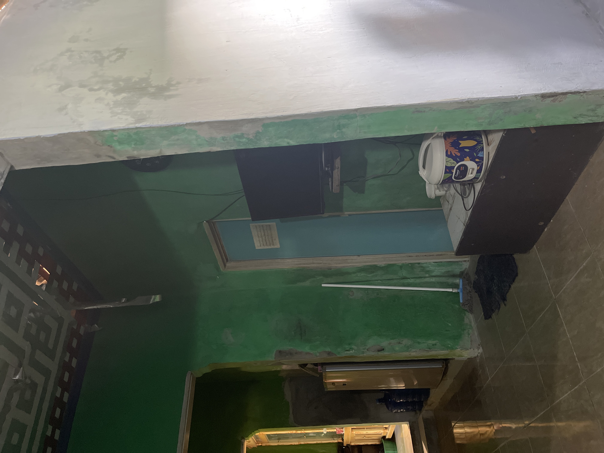 The image depicts an interior scene, likely a room in a residential building in Laban, Indonesia. The walls are painted a vibrant green, contrasting with a white-painted concrete pillar or wall on the right side of the frame. The ceiling shows a geometric pattern and appears to have some damage or incomplete finishing with visible tape.
In the foreground and midground, the tiled floor reflects the ambient light. To the left, a doorway leads to an area with a wooden cabinet, a window with light streaming through, and several large water jugs. A yellow refrigerator is visible next to the cabinet.
Further into the room, a blue door stands open. Beside it, a broom with a blue mop head rests on the floor, next to a dark mat. A flat-screen television is mounted on the wall above a dark wooden cabinet. On top of this cabinet, a white rice cooker with a colorful decorative pattern is placed. Another smaller container is visible next to the rice cooker.
The scene appears to be a domestic living space, possibly a kitchen or living area. The lighting suggests it is daytime, with natural light entering from the window on the left. The overall condition of the room indicates a lived-in environment with some signs of wear or ongoing renovation. There are no people visible in the image.