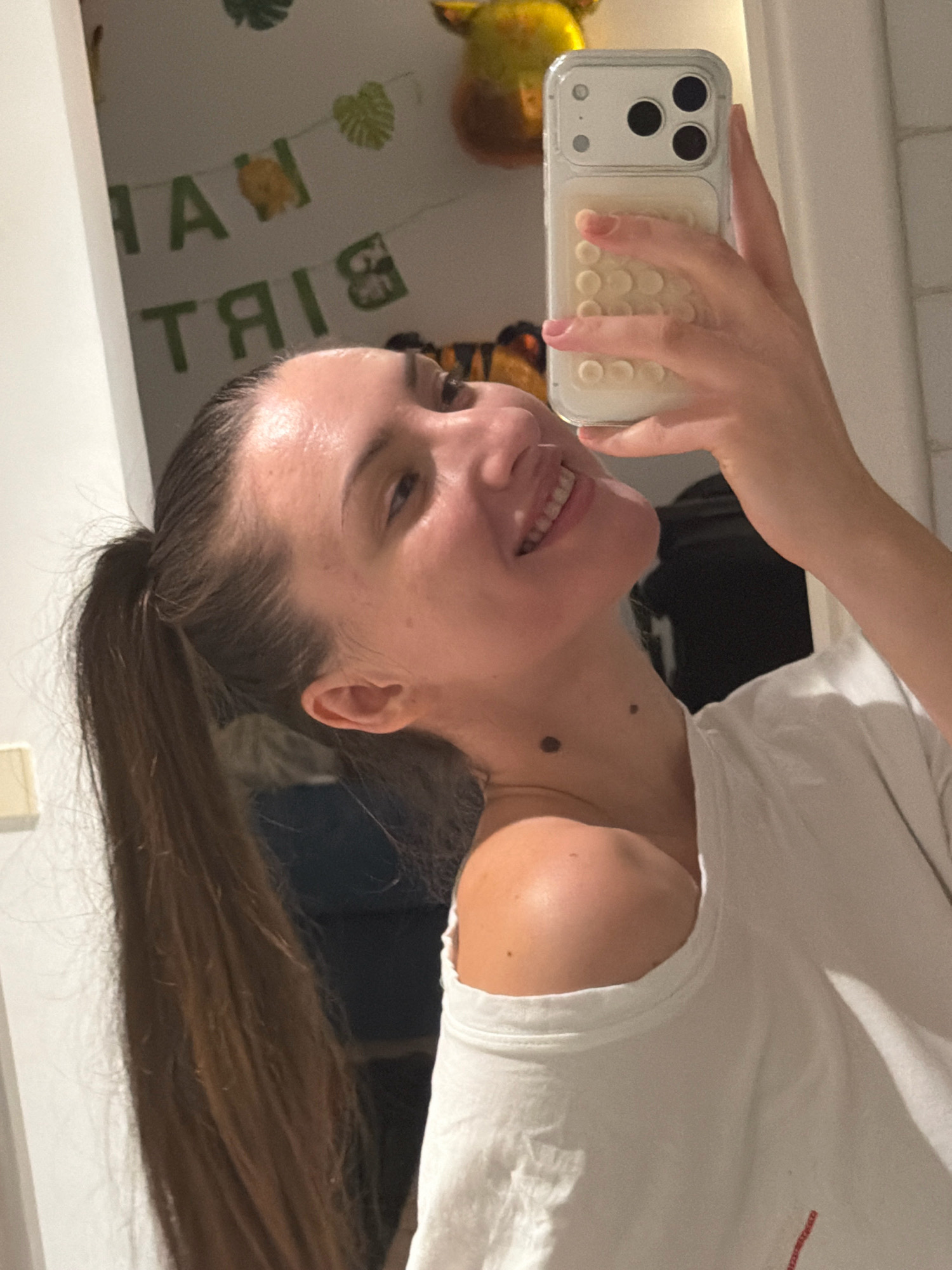 A young woman with brown hair in a ponytail is taking a mirror selfie indoors. She is wearing a white t-shirt and smiling. Behind her, a banner with "HAPPY BIRTHDAY" written on it is visible on the wall, along with some green leafy decorations. The image appears to be taken in a home environment, possibly in Sharm El Sheikh, Egypt.