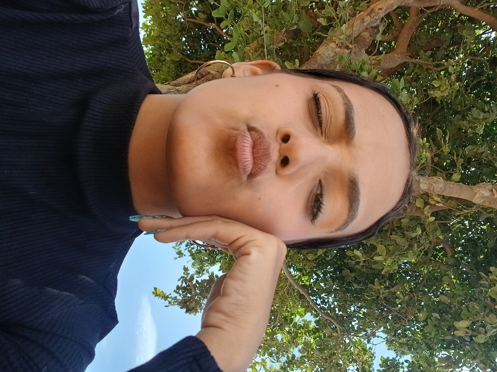 A close-up, low-angle portrait captures a person, likely female, posing outdoors on a sunny day. The subject's face is prominent, rotated 90 degrees clockwise, with dark hair framing their features. Their eyes are closed, and their lips are playfully pursed in a "kissing" or "pout" expression. One hand, featuring blue/teal nail polish, gently rests against their chin and cheek. They wear a dark, ribbed long-sleeved top and a gold hoop earring is visible in one ear.
The background is split between a clear, bright blue sky and a dense canopy of vibrant green tree leaves and branches, indicating a natural, outdoor setting, possibly under a large tree. The bright lighting suggests it is daytime, likely a clear and sunny morning or afternoon. The low camera perspective creates an upward-looking view. The pose conveys a playful or serene mood. No text is visible in the image. The location context is Altagracia, Venezuela.