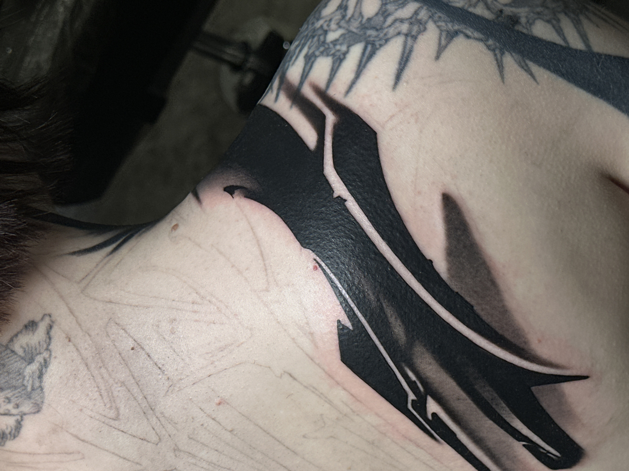 The image presents a close-up view of a person's upper body, focusing on their shoulder and chest area. A large, bold blackwork tattoo is prominently featured, characterized by abstract, angular shapes, intense black ink, and intricate negative space lines that create depth. The fresh appearance of the tattoo, along with some visible redness and irritation on the surrounding light-toned skin, suggests that it is either actively being worked on or has just been completed.
Above this new blackwork, on the shoulder/collarbone, an older, faded tattoo depicting what appears to be a crown of thorns or a similar thorny motif is visible in grey tones. To the left, near the neck and upper back, another existing tattoo with a lighter, possibly mushroom-themed design is partially seen, along with the person's dark hair. Faint stencil lines for future tattoo work are evident on the un-tattooed skin, outlining the intended expansion of the design with more geometric or abstract patterns.
The scene is an indoor setting, likely a tattoo studio, with a dark, out-of-focus background. There is no visible text. The location context provided is Moscow, Russia.