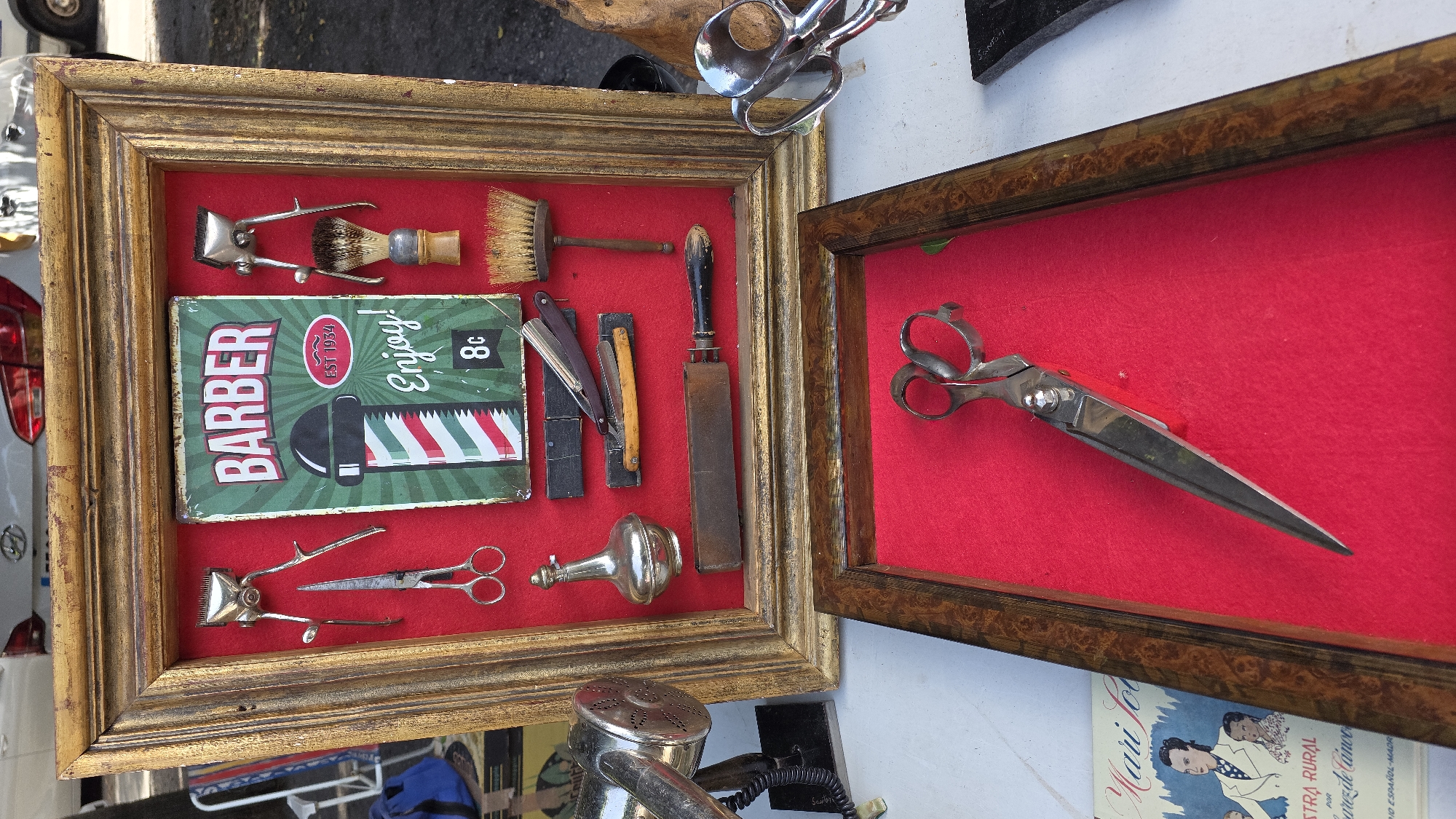 The image depicts a stall at an outdoor market, likely a flea market or antique fair, with items displayed for sale. The primary focus is a framed collection of vintage barber tools. The collection is mounted on a red felt background within a golden, ornate frame. Items visible include a manual hair clipper, scissors, a small glass bottle, a straight razor with a wooden handle, a sharpening strop, and two shaving brushes. A vintage "Barber" sign, featuring a barber pole design and the text "EST 1934 Enjoy!", is also prominently displayed.
Below this framed display, a separate, smaller wooden frame with a red felt interior holds a pair of large, antique scissors. To the left of the framed items, partially visible, is a vintage silver hair dryer. Further to the left and bottom, a booklet or small book with illustrations of people and text in Spanish, possibly related to rural life or a magazine, is visible. The background suggests an outdoor setting with a vehicle and trees. The overall impression is one of vintage Americana or historical grooming tools.
While the image itself does not provide explicit geographical markers for Almuñécar, Spain, such markets are common in tourist towns and historical centers. If this stall is indeed in Almuñécar, it would likely be situated in or near the town's historic center, such as the Plaza del Ayuntamiento or along the main streets leading to the beach or the Castillo de San Miguel. The presence of vintage items and crafts is typical of such locations.