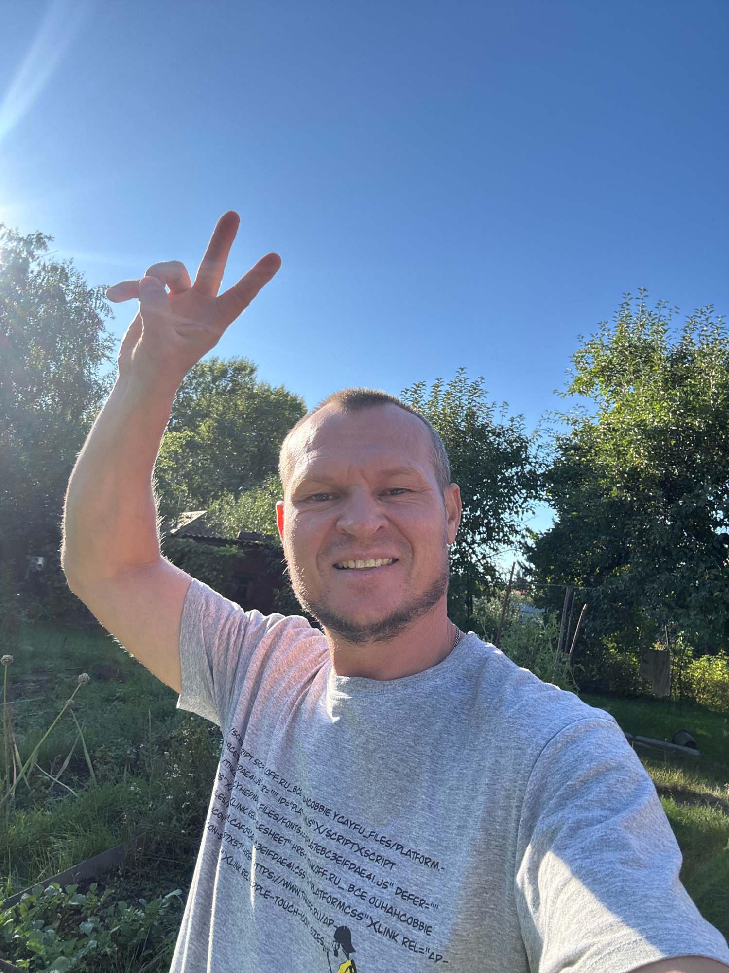 A middle-aged man with a beard and short hair is taking a selfie outdoors. He is wearing a gray t-shirt with text printed on it. His right arm is raised, and he is making a peace sign with his fingers. The background shows a clear blue sky, green trees, and some vegetation. The lighting suggests it is daytime, possibly late afternoon, due to the strong sunlight and shadows. The location is the city of Novo-İavlenka, Kazakhstan.