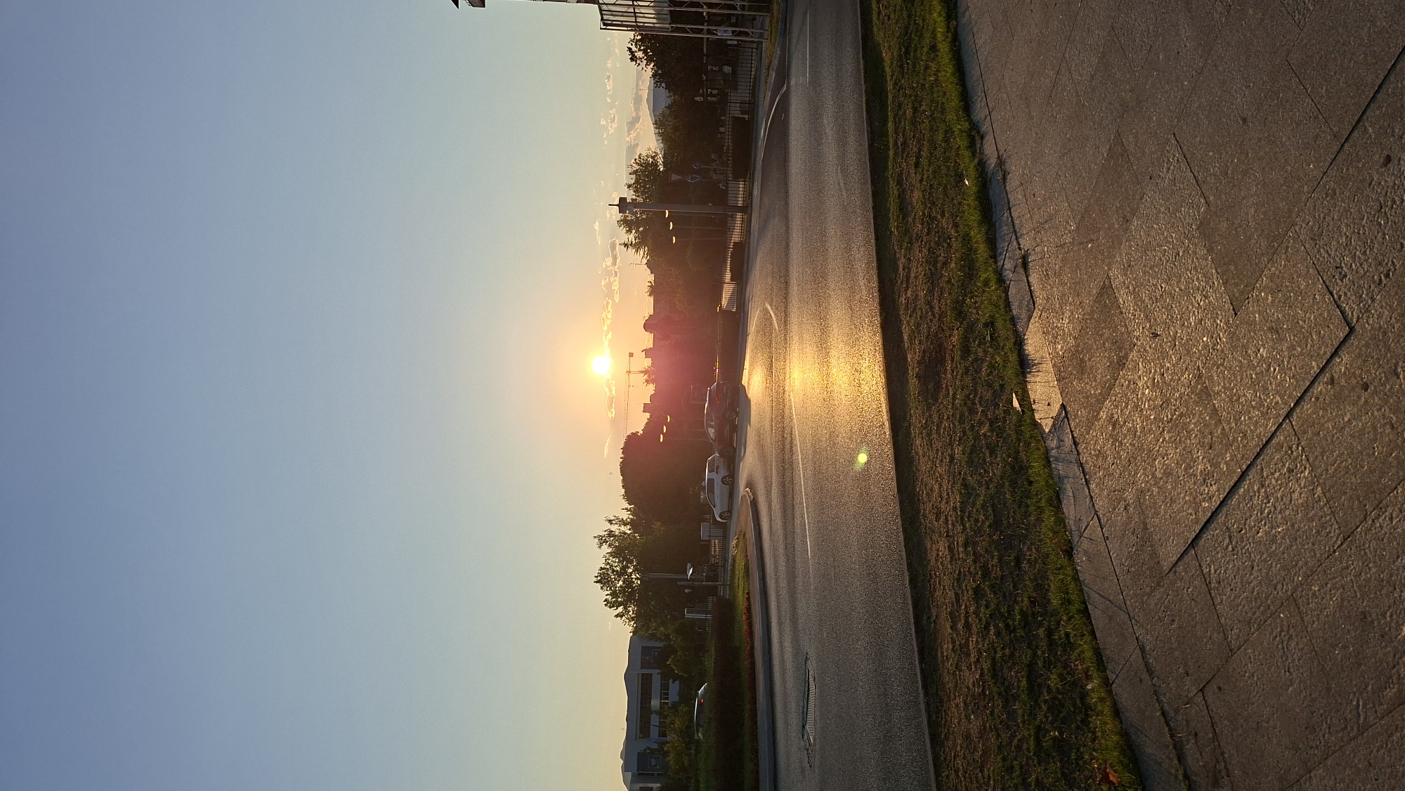 The image captures a sunset in Podgorica, Montenegro, likely in a more modern, developed area of the city. The sun, a bright orb with a reddish-orange hue, is setting low on the horizon, casting a warm, golden glow across the scene. The sky is a clear gradient of pale blue at the top, transitioning to a softer, warmer tone near the horizon, with a few wispy clouds.
In the foreground, a paved pedestrian walkway is visible, made of large, light-colored stone tiles arranged in a pattern. Adjacent to the walkway is a strip of green grass, with a few scattered dry leaves. Beyond the grass, a two-lane road curves through the scene. The asphalt of the road is wet, reflecting the intense golden light of the sunset, creating a luminous sheen.
Several cars are present on the road. On the left side, a white car is turning or driving on a circular junction area with a green, landscaped median. Further down the road, two cars, a darker colored sedan and a white hatchback, are parked or stopped, bathed in the sunset's light. In the background, a cluster of trees with dark green foliage lines the edge of the road. Beyond the trees, buildings are visible, including a multi-story building with a modern facade on the left and other structures with rooftops. A construction crane is also visible in the distance, silhouetted against the bright sky. Streetlights and traffic signs are scattered along the roadside. The overall impression is that of an urban or suburban environment during a calm, picturesque sunset.